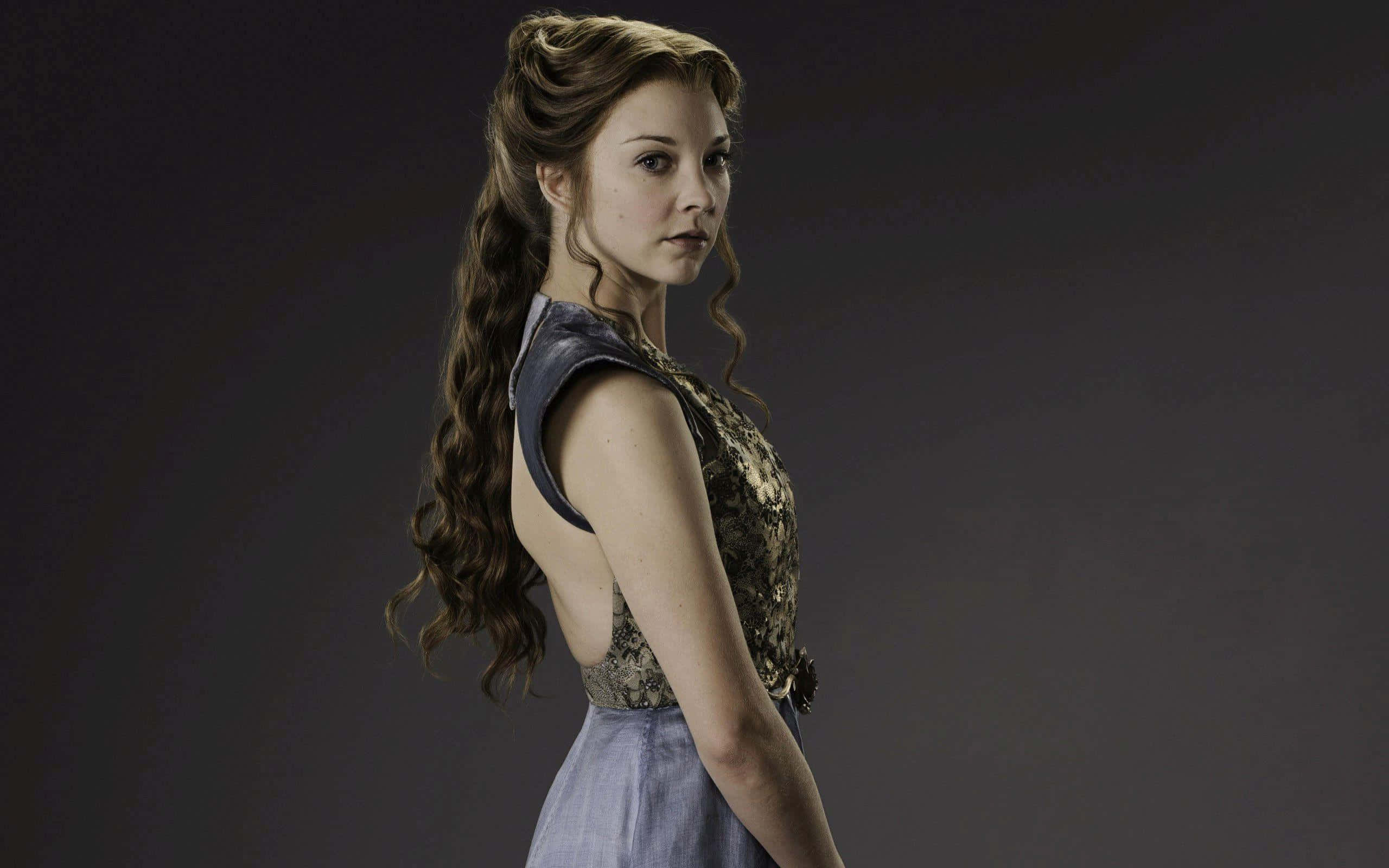 natalie dormer 桌布 壁紙 — 一位擁有長波浪棕色頭髮的女性，穿著華麗的連衣裙，在深灰色背景上轉頭越過肩膀。