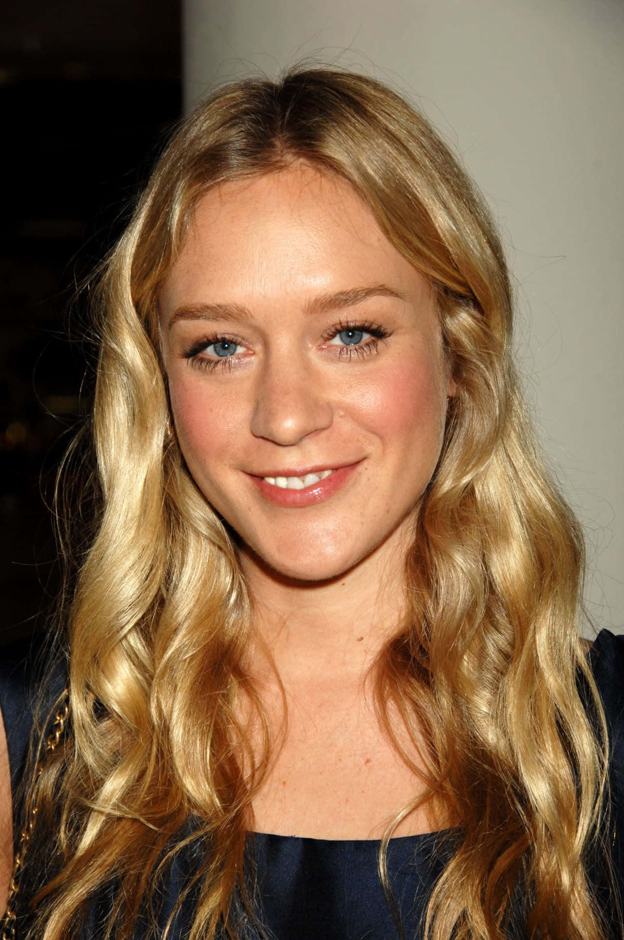 Chloë Sevigny 桌布 壁紙 — 一位擁有長波浪金髮和明亮藍眼睛的女性特寫肖像，溫暖地對著鏡頭微笑。