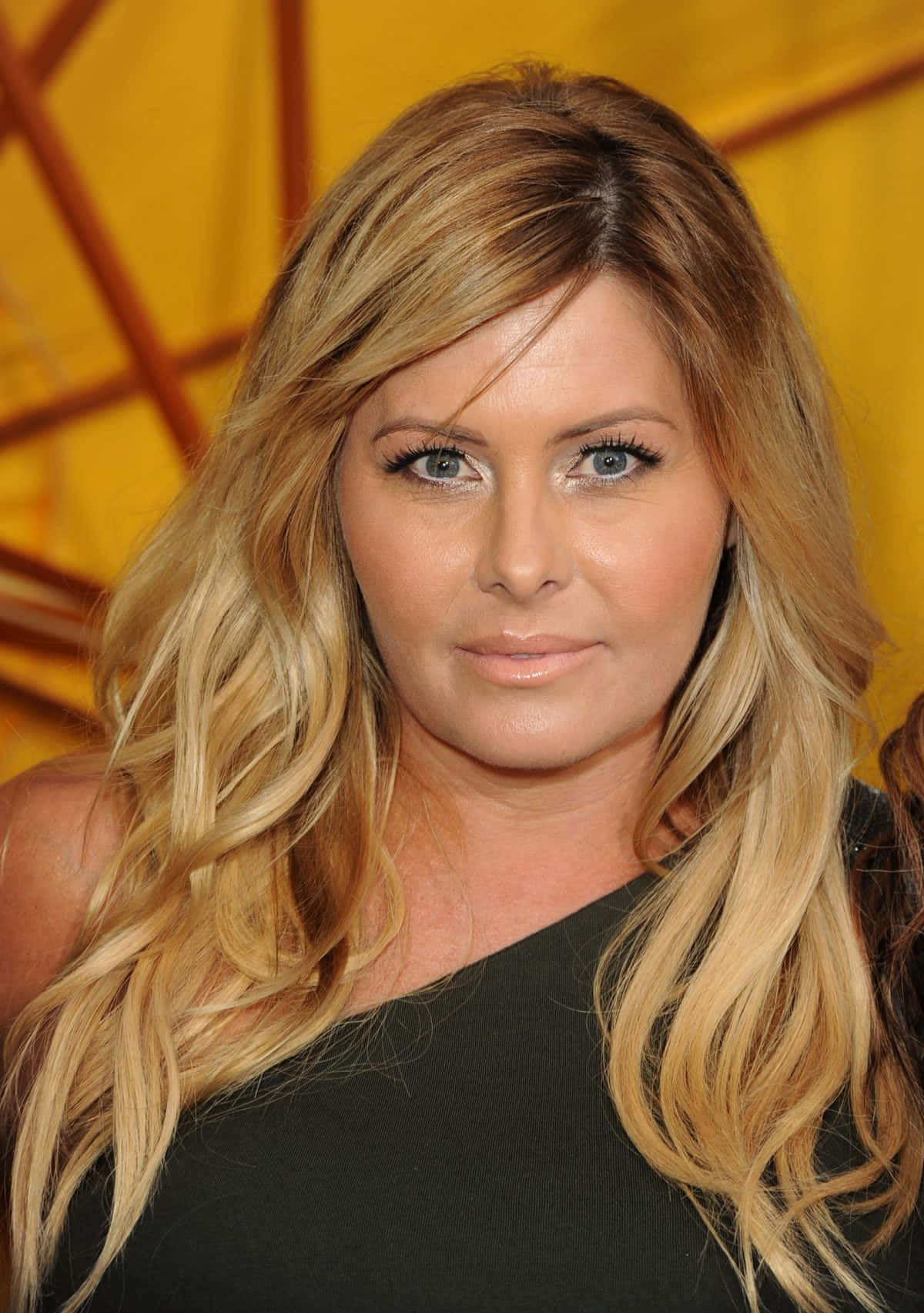 Nicole Eggert 桌布 壁紙 — 一位擁有長波浪金髮和藍眼睛的女性，身穿黑色上衣，以溫暖的黃色幾何背景為襯托的特寫肖像。