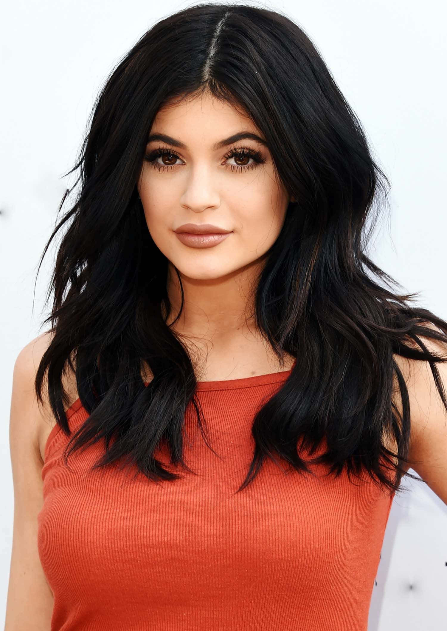 Kylie Jenner 桌布 壁紙 — 一位擁有長深色波浪髮和精緻妝容的女性，在純白色背景前穿著合身的橙色羅紋上衣。