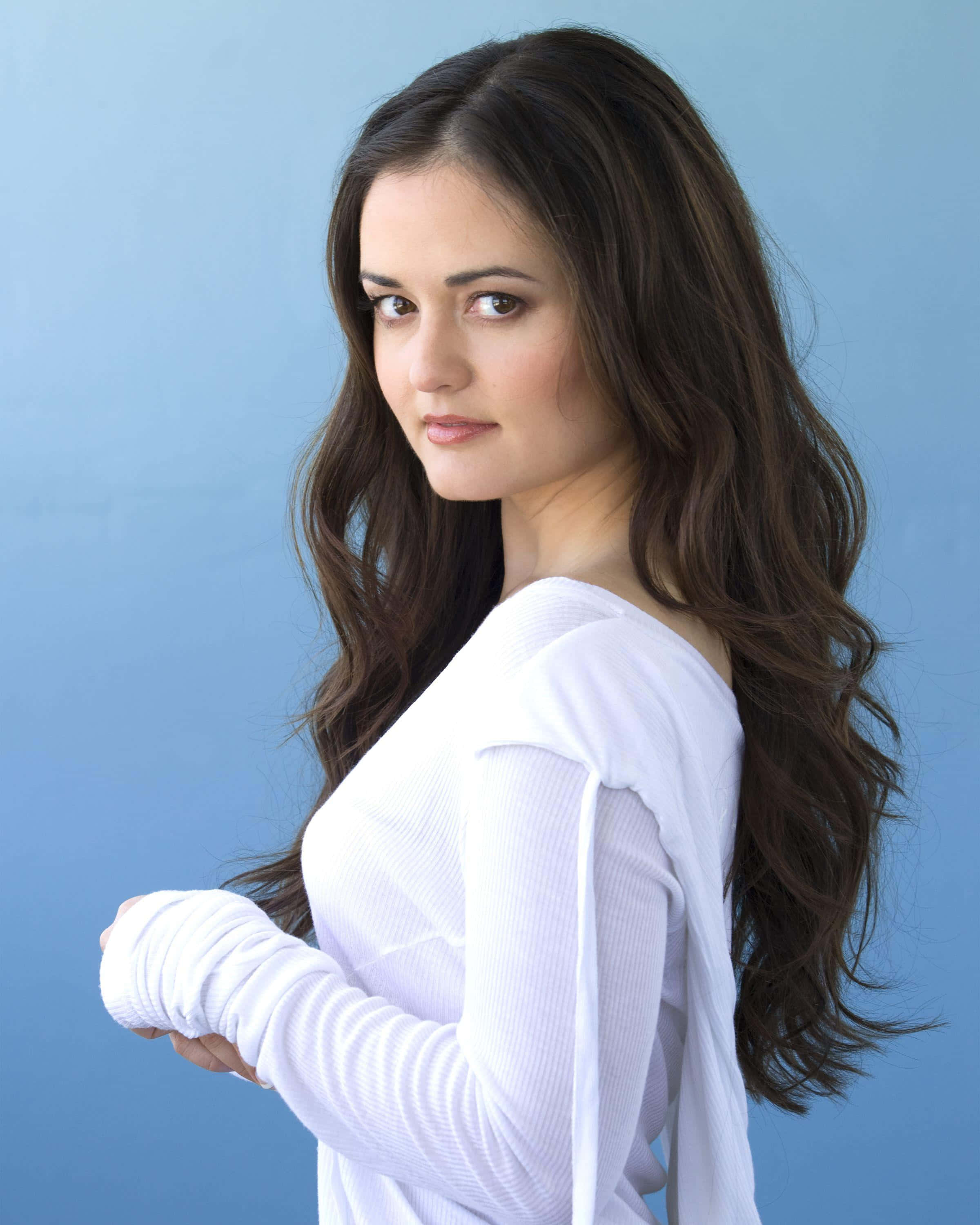 Danica Mckellar 桌布 壁紙 — 一位擁有長深色波浪髮的女性，在純藍色攝影棚背景前穿著白色羅紋上衣。