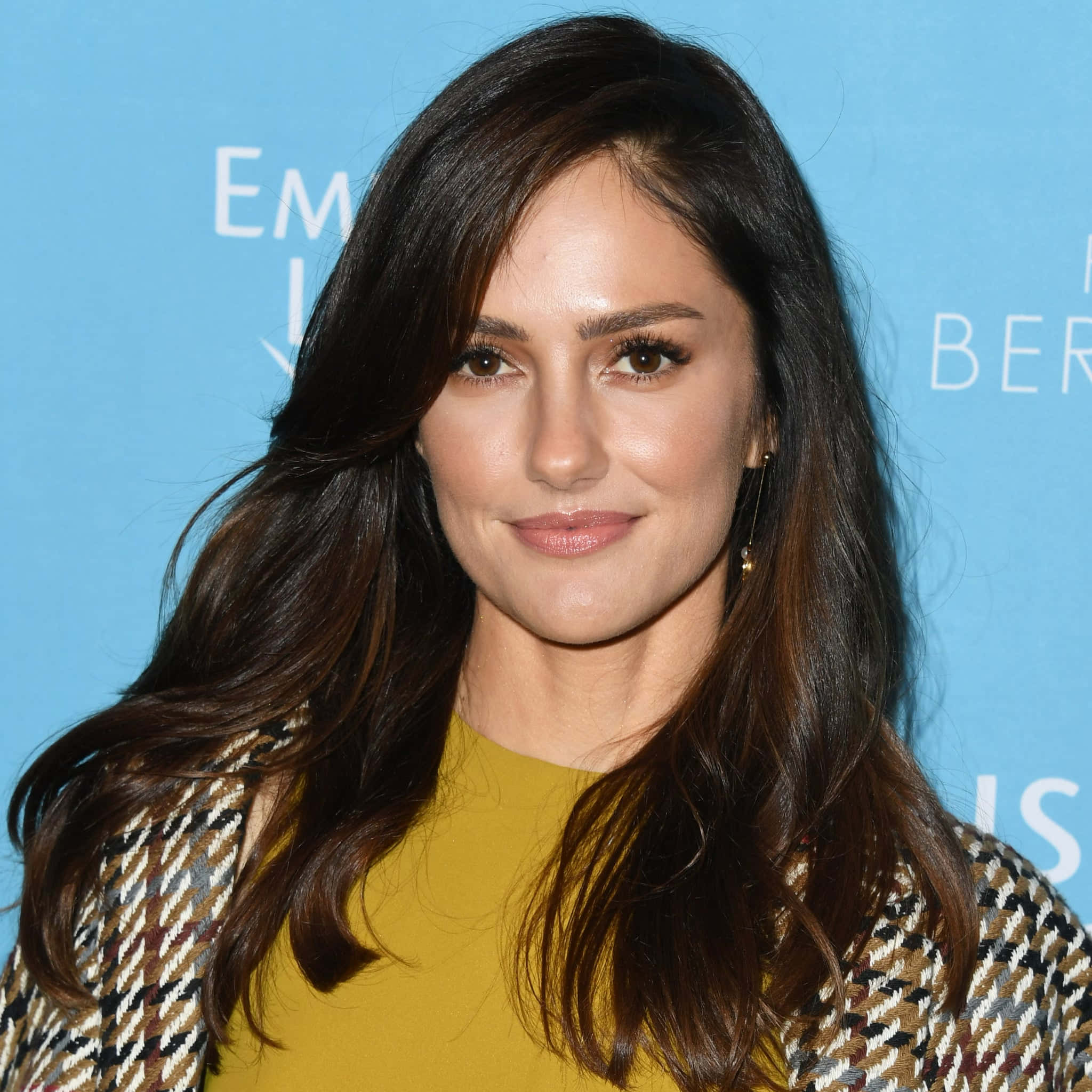 minka kelly 桌布 壁紙 — 一位擁有長深色波浪髮的女性，穿著芥末色上衣和圖案外套，在藍色背景上帶有白色文字，溫柔地微笑。