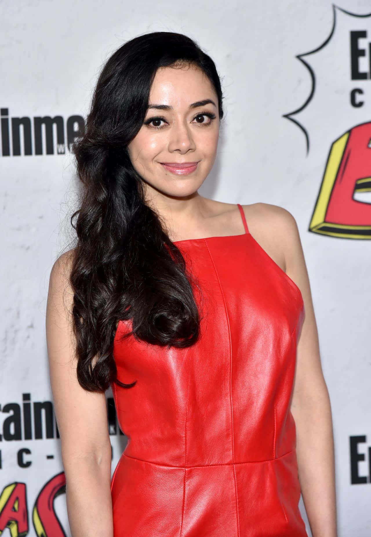 Aimee Garcia 桌布 壁紙 — 一位擁有長深色波浪髮的女性，穿著鮮紅色皮裙，在漫畫活動背景下微笑。