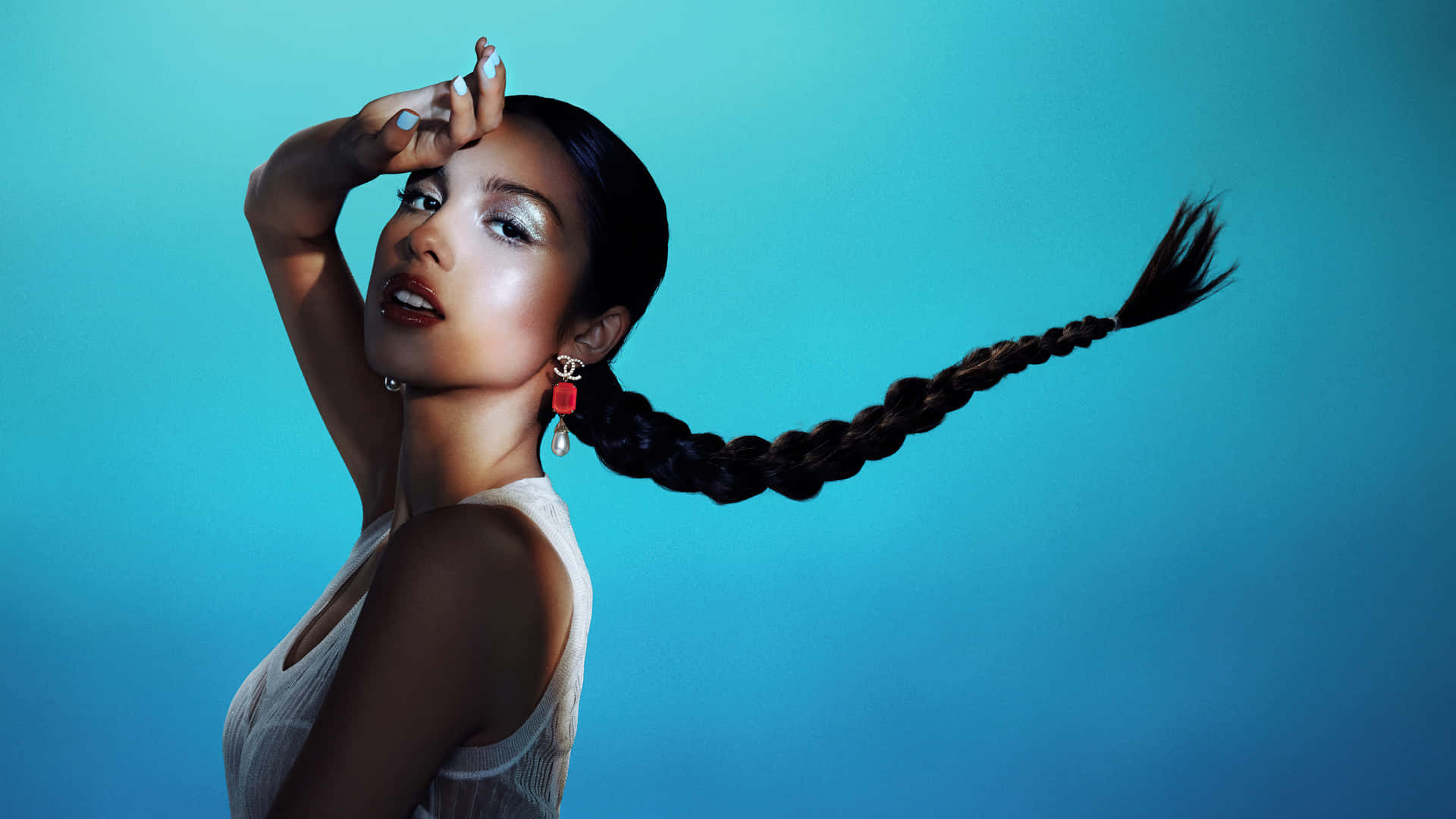 Olivia Rodrigo 桌布 壁紙 — 一位擁有長辮子髮型的女性在充滿活力的藍色背景前擺姿勢，手靠近額頭，穿著白色上衣和紅色耳環。