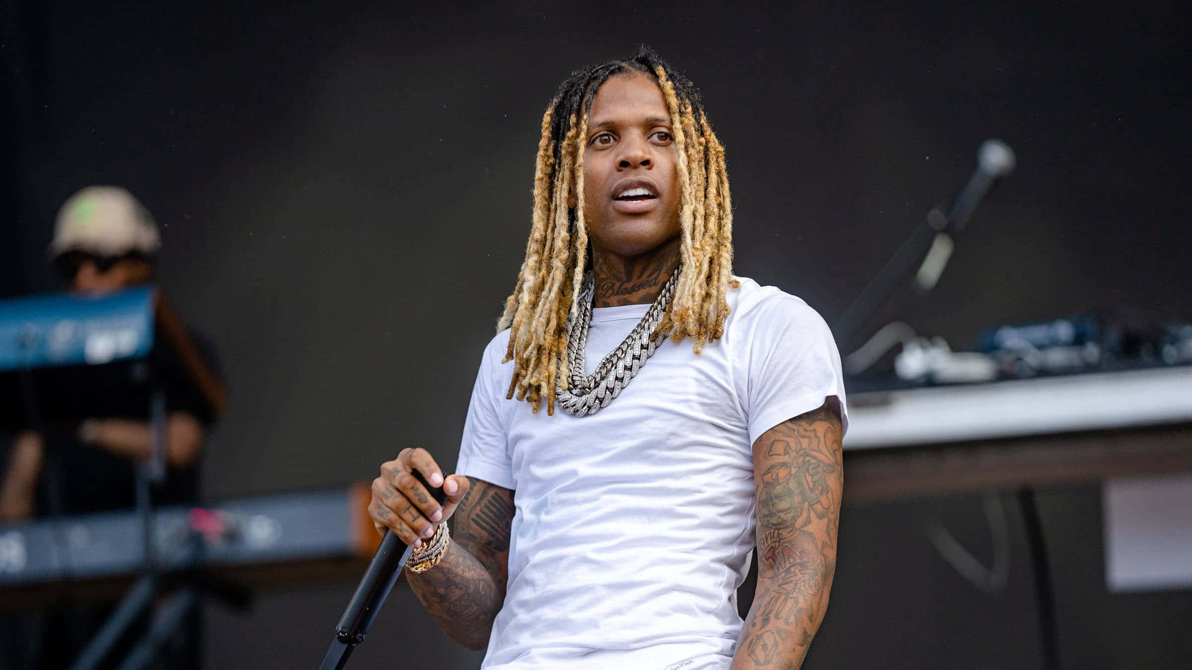 lil durk 桌布 壁紙 — 一位擁有長金髮辮和紋身的說唱歌手在舞台上表演，手持麥克風，身穿白色T恤和層疊鏈條。