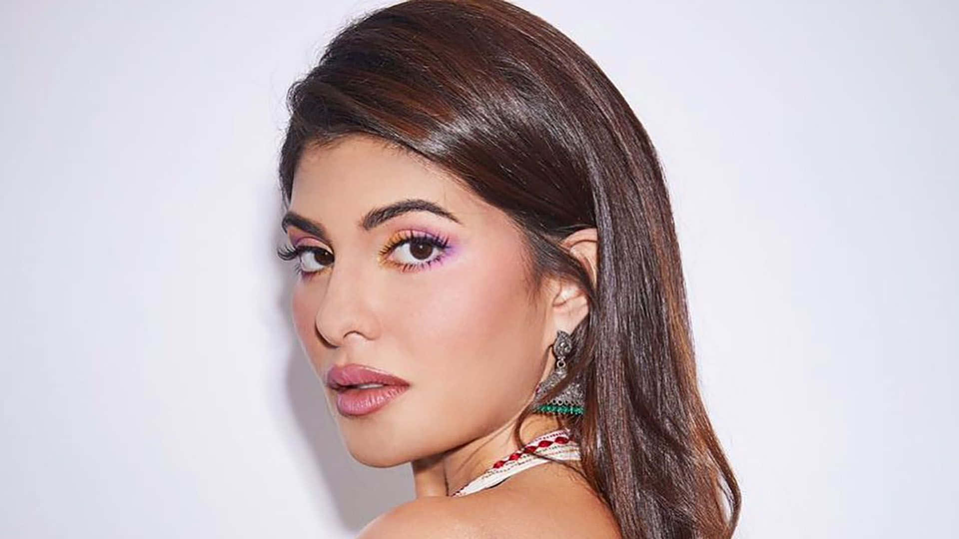 Jacqueline Fernandez 桌布 壁紙 — 一位擁有長長棕色頭髮和鮮豔紫色眼影的女性，在純白色背景上回頭凝視。