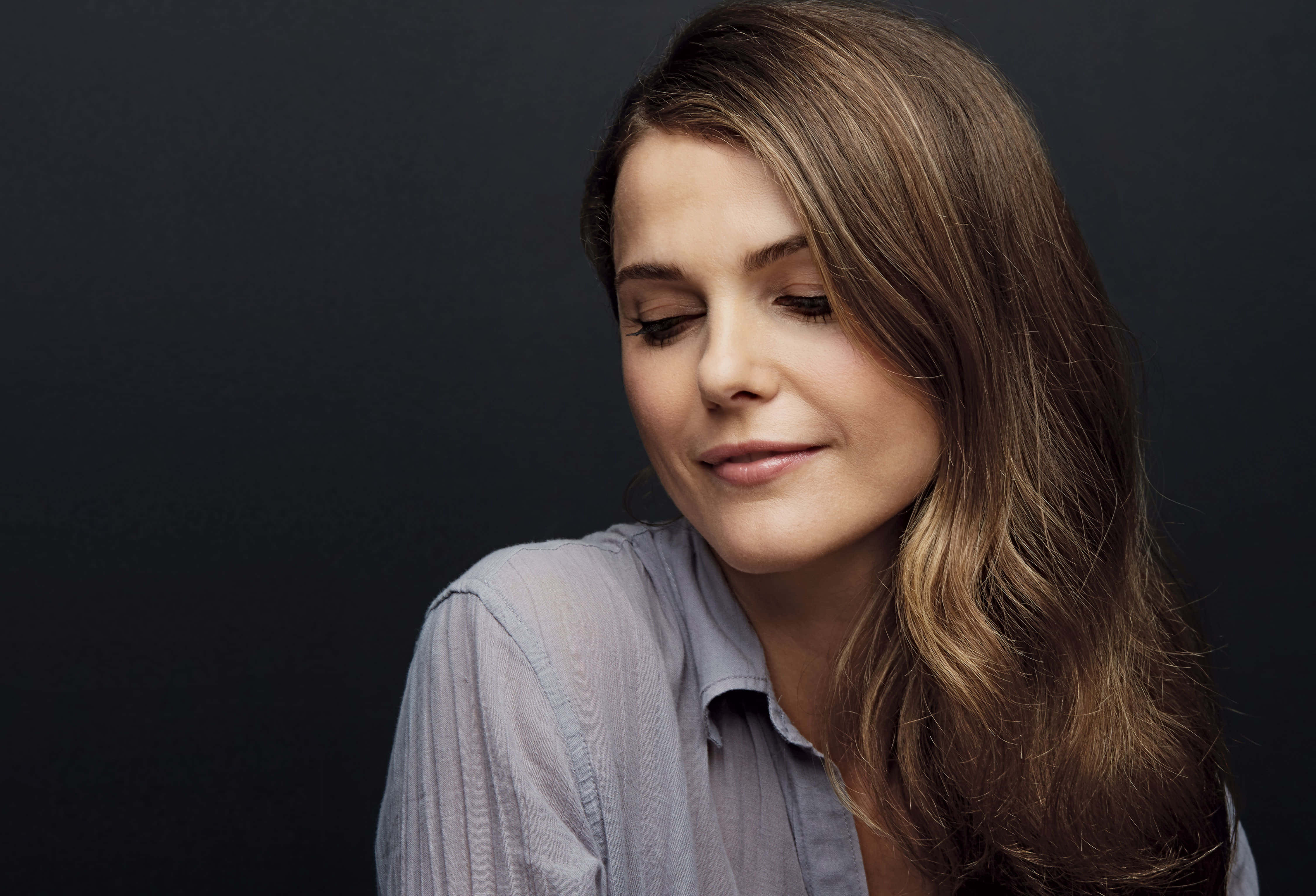 keri russell 桌布 壁紙 — 一位擁有長長棕色頭髮的女性在深色背景下低頭微笑。