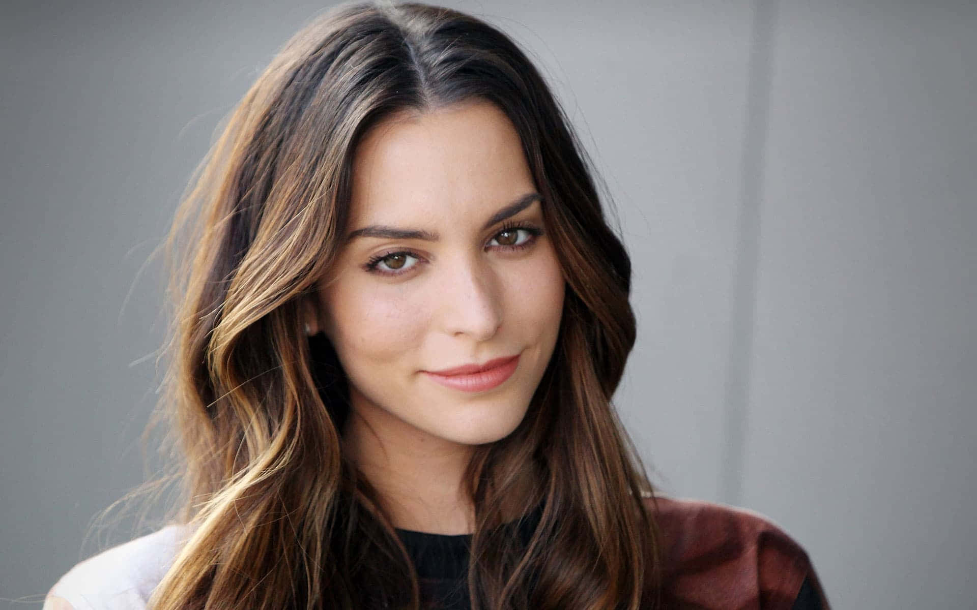 Genesis Rodriguez 桌布 壁紙 — 一位擁有長長棕色頭髮的女性在純灰色背景前溫柔地微笑。
