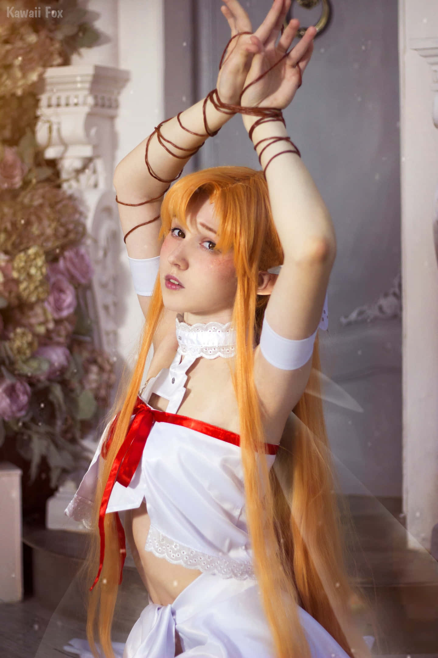 Sword Art Online Cosplay 桌布 壁紙 — 一位擁有長長橙色頭髮的cosplayer，身穿帶有紅色蝴蝶結的白色連衣裙，在花卉和建築背景前優雅地舉起雙臂擺姿勢。