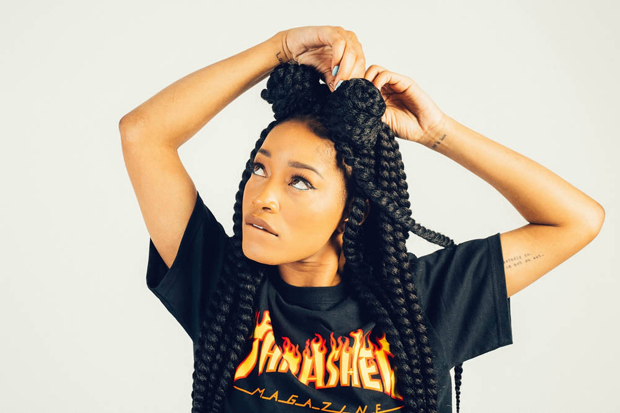 Keke Palmer Thrasher 壁紙 — 一位擁有長長黑色辮子、梳成兩個髮髻的女性，在純白背景下向上看，同時整理頭髮。