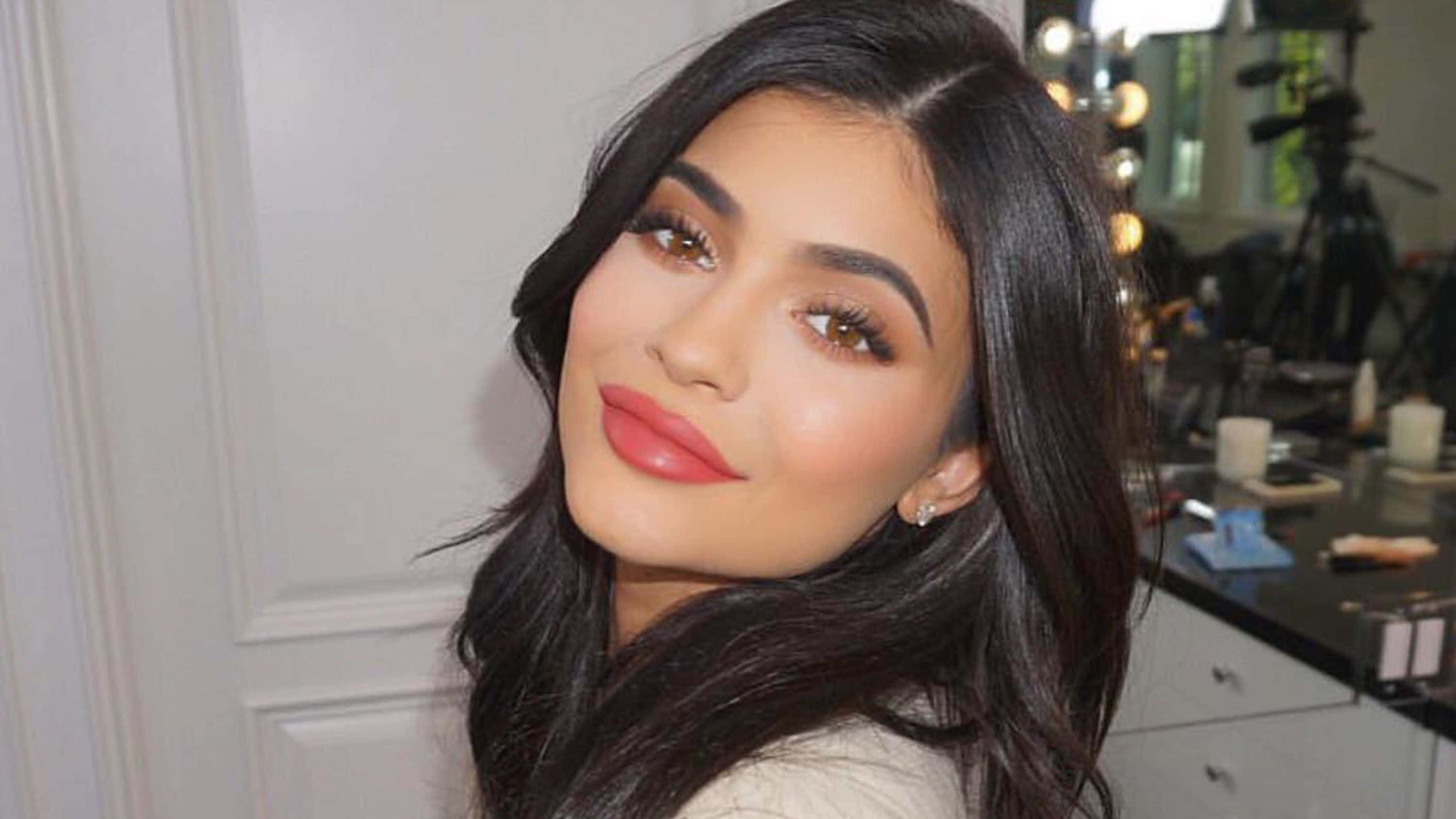 Kylie Jenner 桌布 壁紙 — 一位擁有長黑髮和大膽紅唇的魅力女性，在光線充足的化妝室中溫柔微笑。