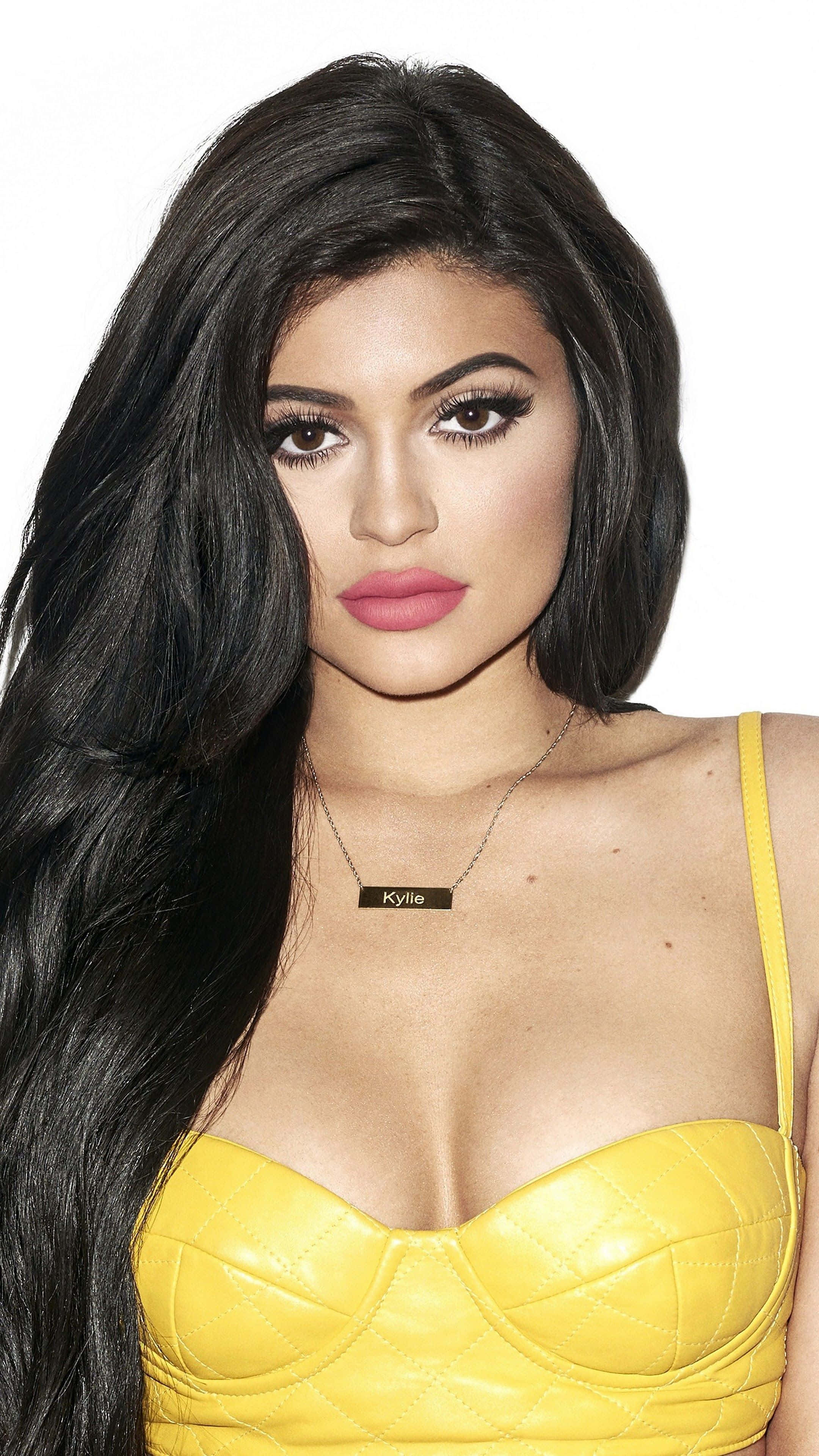 Kylie Jenner 桌布 壁紙 — 一位擁有長黑髮和濃妝的女性，在純白背景前穿著亮黃色絎縫上衣。