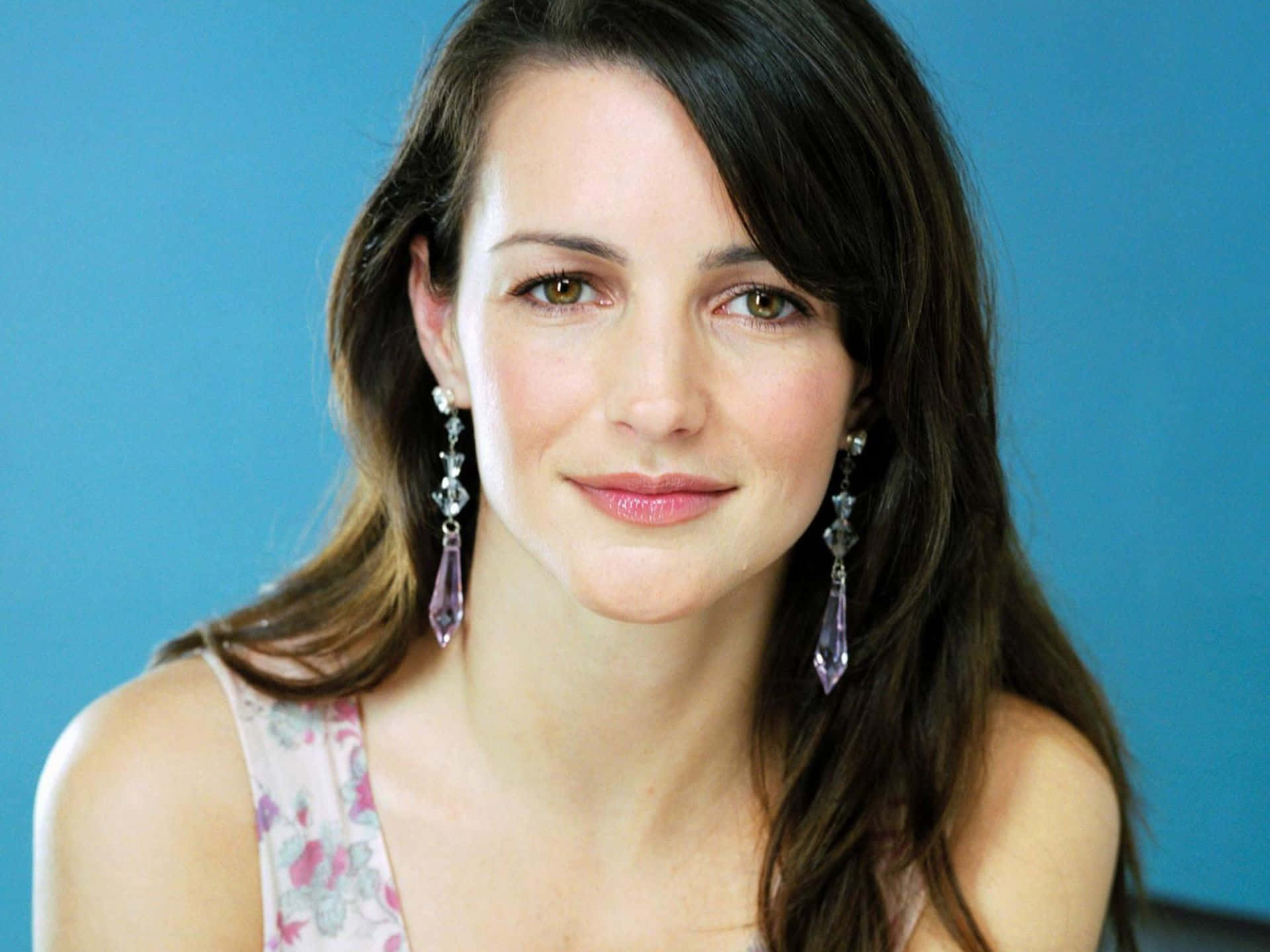 kristin davis 桌布 壁紙 — 一位擁有長黑髮和紫色垂墜耳環的女性在純藍色背景前溫柔微笑。