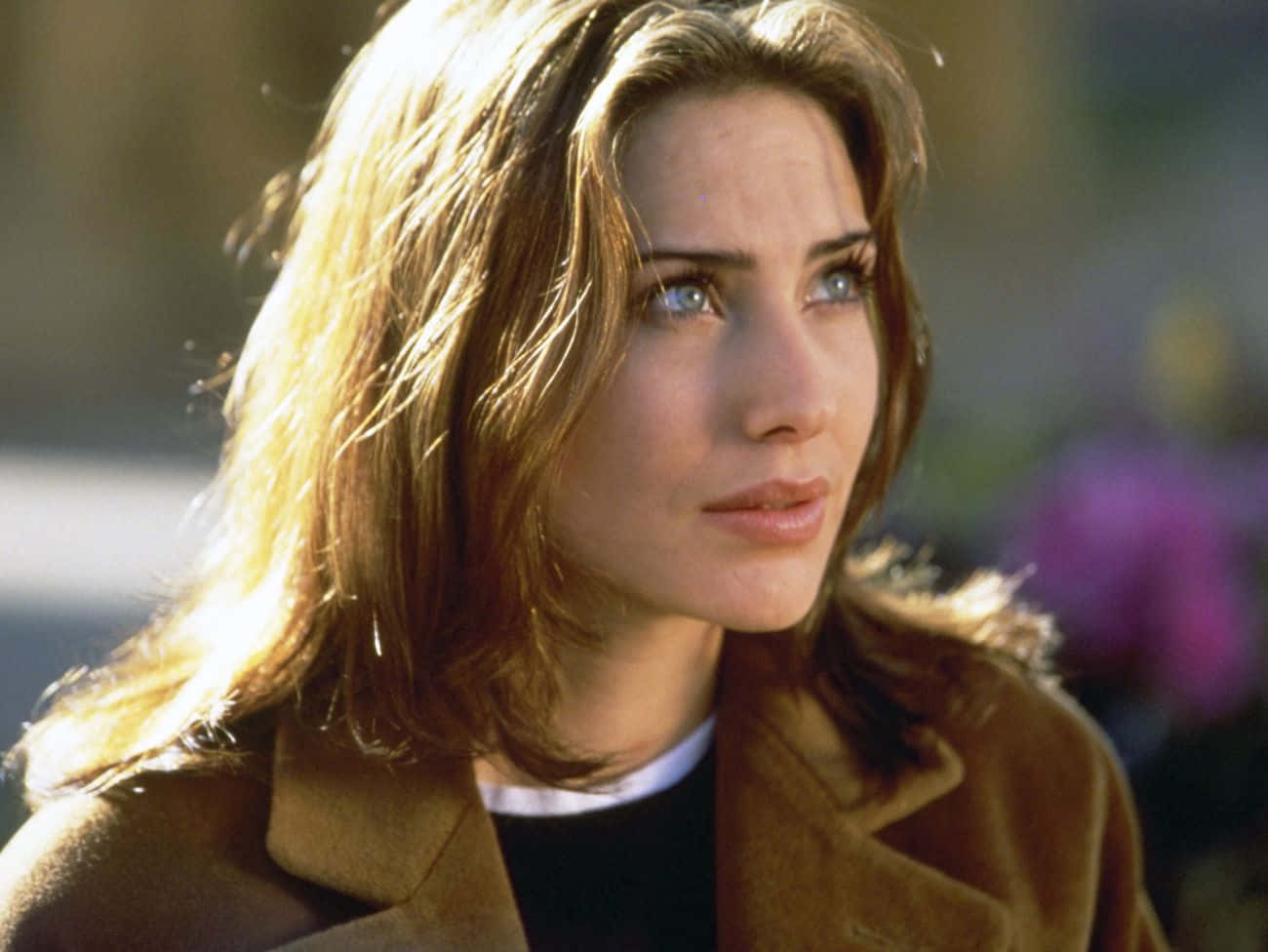 Claire Forlani 桌布 壁紙 — 一位擁有陽光照射的棕色頭髮和藍色眼睛的女性，穿著棕色外套，若有所思地望向戶外。