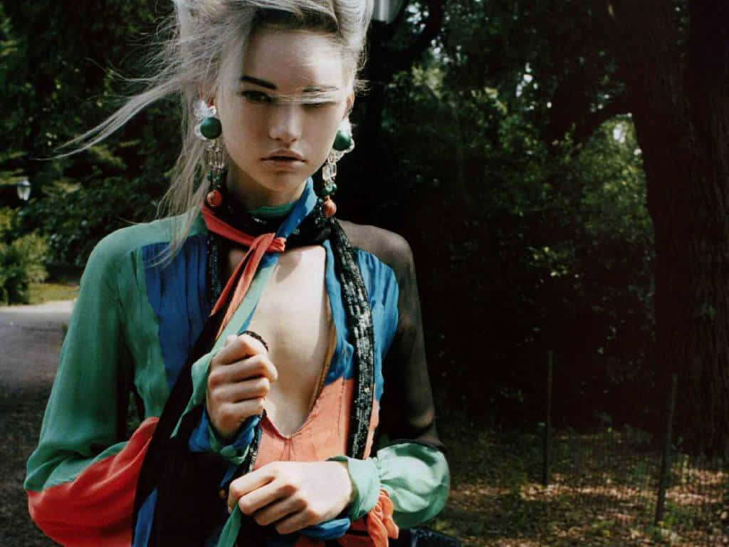 Gemma Ward 桌布 壁紙 — 一位擁有隨風飄逸銀髮和醒目綠色耳環的女性，站在陽光普照的森林中，穿著色彩繽紛的層次服裝。