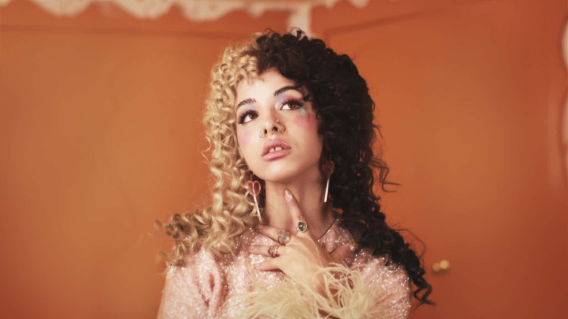 Melanie Martinez 桌布 壁紙 — 一位擁有雙色捲髮和閃亮化妝的女性，在溫暖的橙色牆壁前向上凝視。