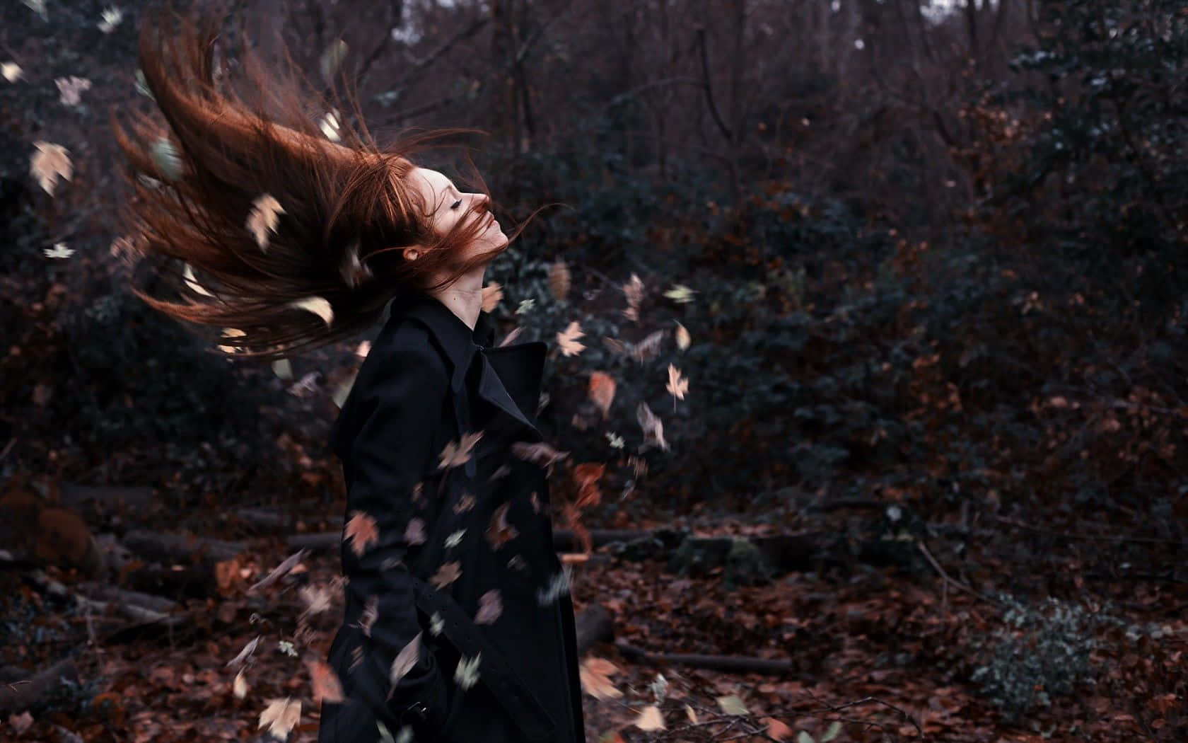 Autumn Wind 桌布 壁紙 — 一位擁有飄逸紅髮的女性在黑暗的森林中拋灑秋葉，她的外套隨風飄揚，風穿過樹林。