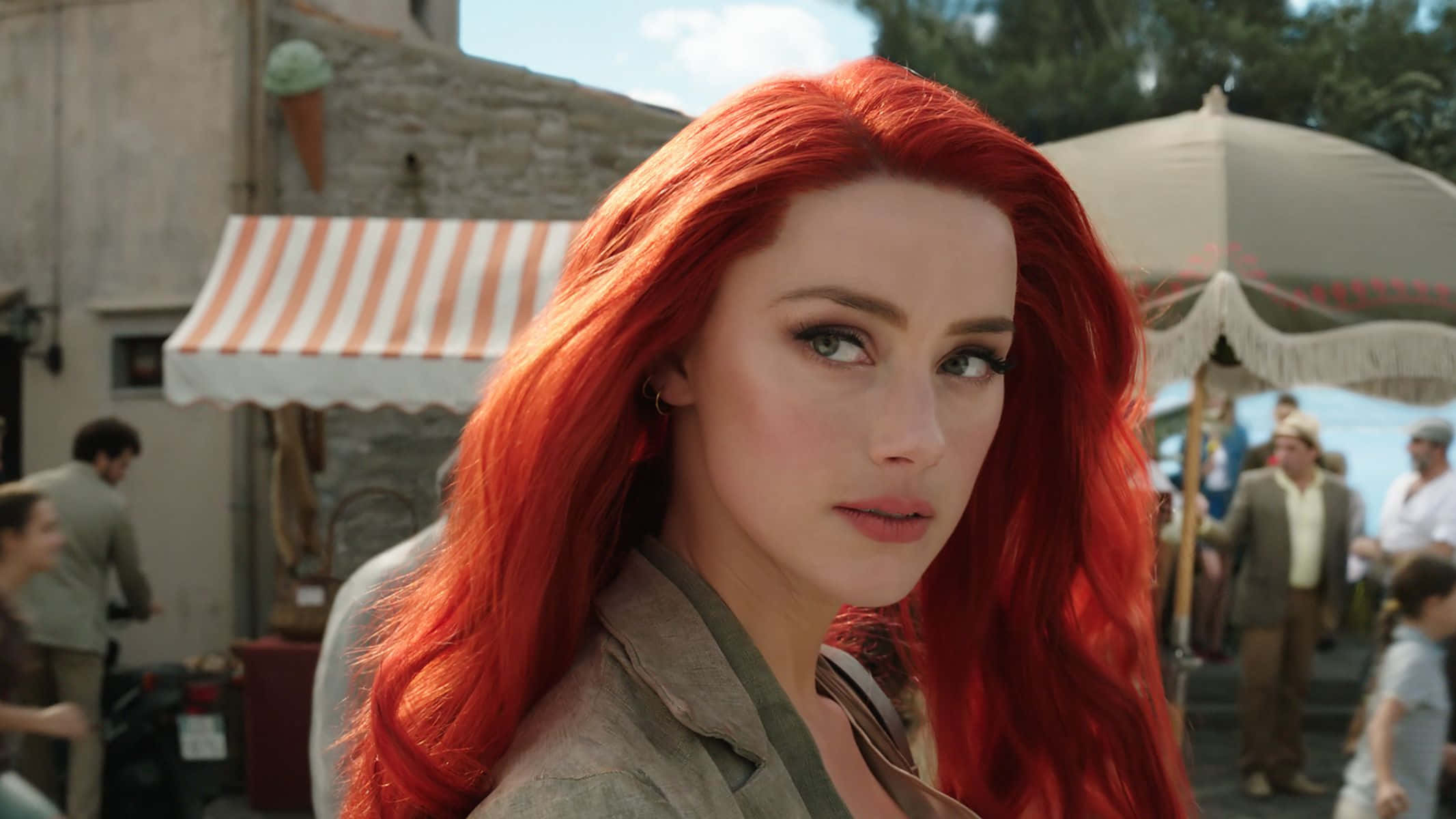 Amber Heard 桌布 壁紙 — 一位擁有鮮豔紅髮的女性在陽光普照的歐洲街景中若有所思地凝視，街道上有條紋遮陽篷和行人。