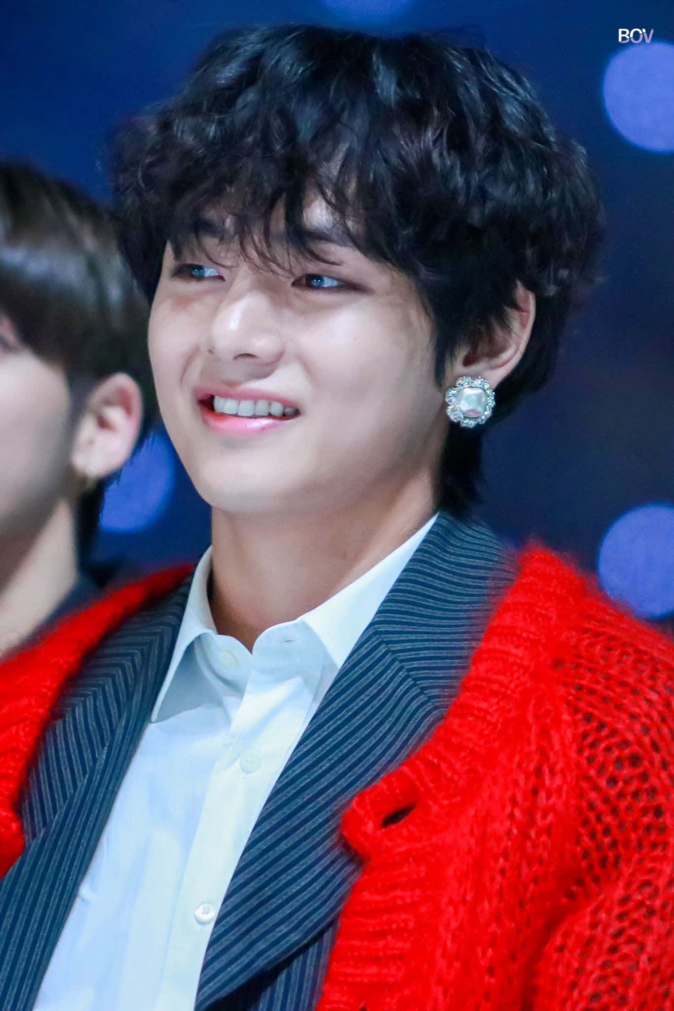 Taehyung Cute 桌布 壁紙 — 一位擁有黑色捲髮的年輕男子，穿著紅色針織開襟衫搭配條紋西裝和閃亮耳環，露出溫暖的笑容。