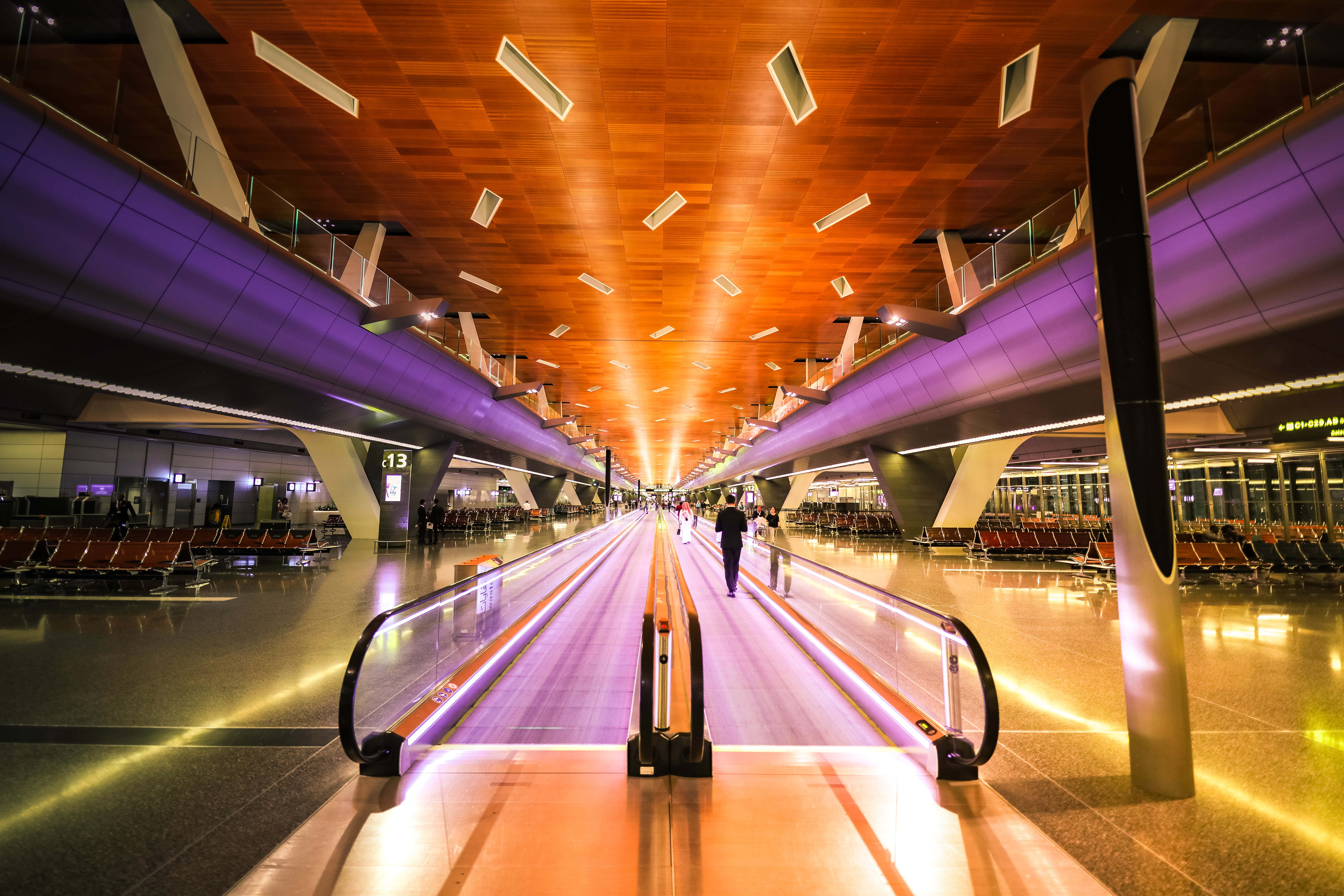 Airport 桌布 壁紙 — 一位旅客在現代機場客運大樓內，沿著發光的紫色自動行人道行走，天花採用溫暖的木質設計，並配以環境照明。