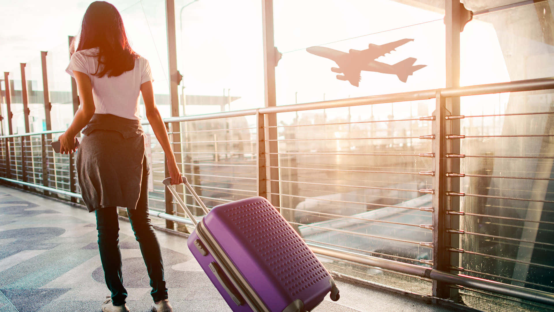 Purple Travel Bag 桌布 壁紙 — 一位旅行者拖著紫色行李箱穿過機場客運大樓，此時正值黃金時段，一架飛機從頭頂飛過。