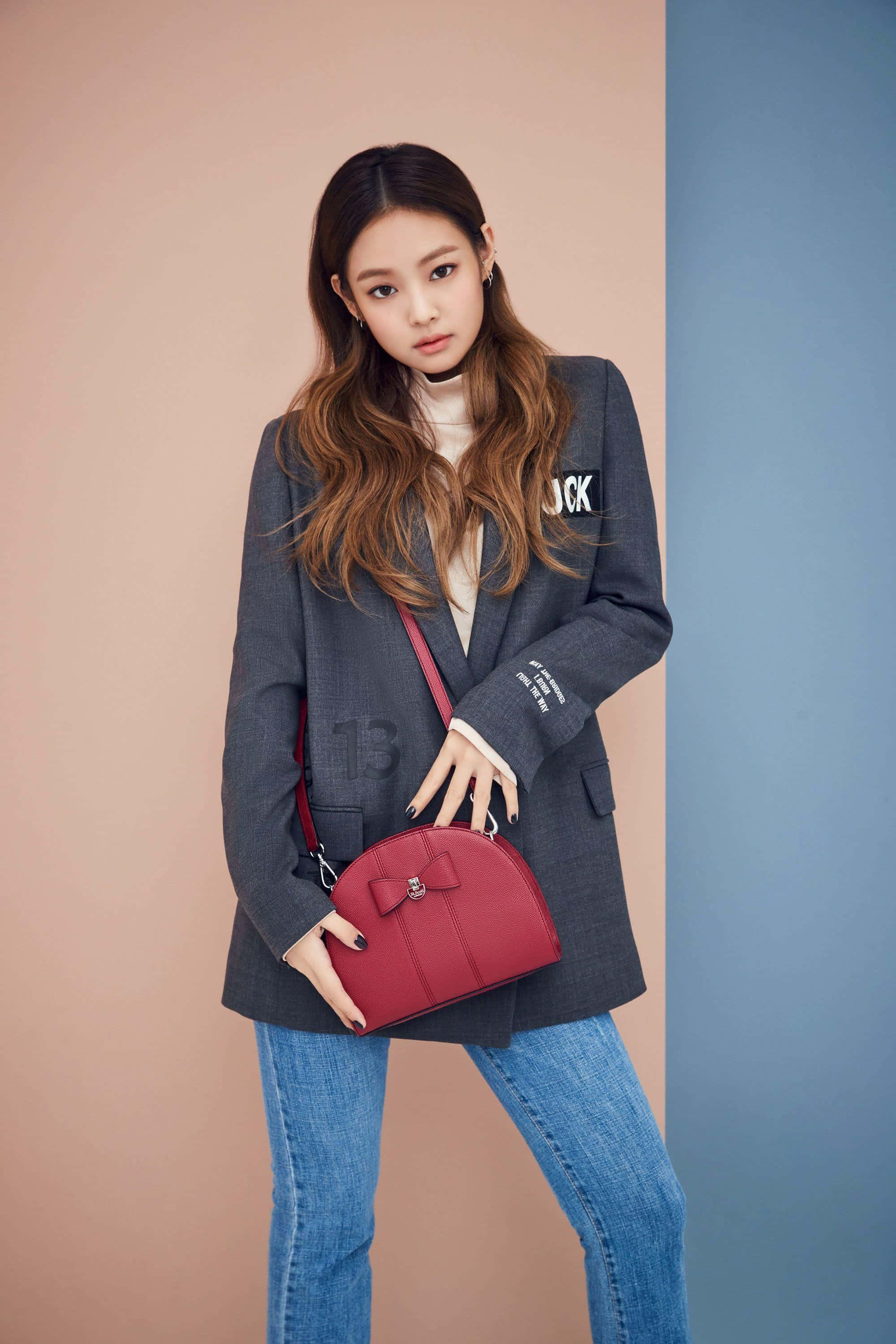 Jennie Kim 桌布 壁紙 — 一位時尚女性穿著灰色西裝外套和藍色牛仔褲，在粉紅色和藍色分割的攝影棚背景前拿著一個紅色手袋。