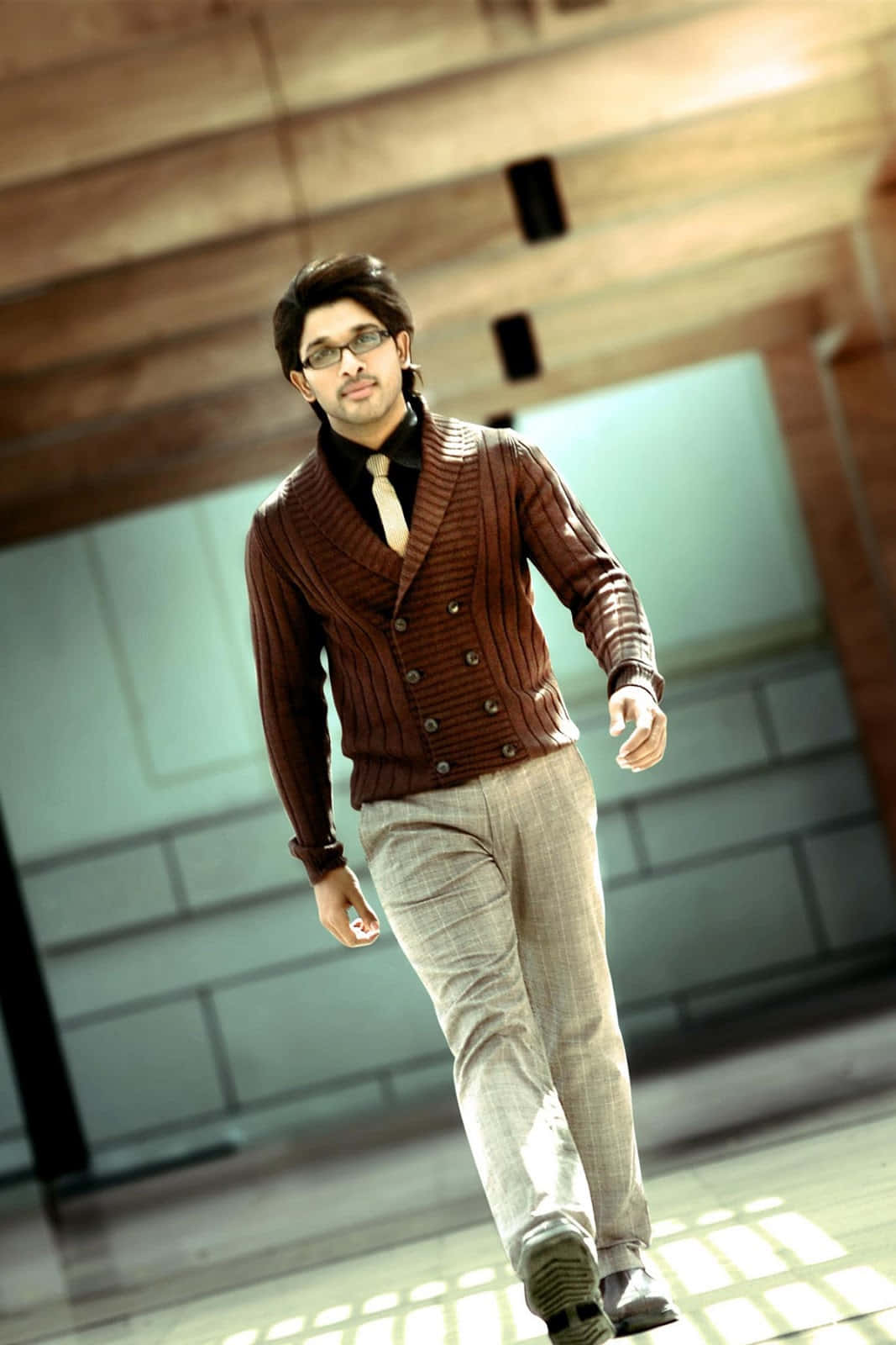 Allu Arjun 桌布 壁紙 — 一位時尚男士穿著棕色羅紋開襟毛衣和眼鏡，自信地走過現代木質和玻璃室內空間。