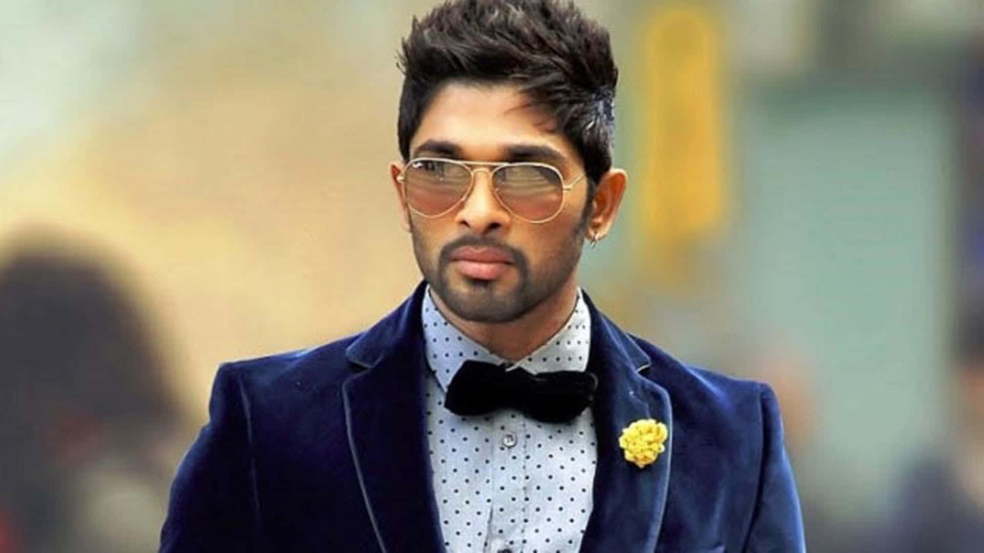 Allu Arjun 桌布 壁紙 — 一位時尚男士身穿海軍藍天鵝絨西裝和領結，戴著飛行員太陽眼鏡和黃色襟花，自信地擺姿勢。