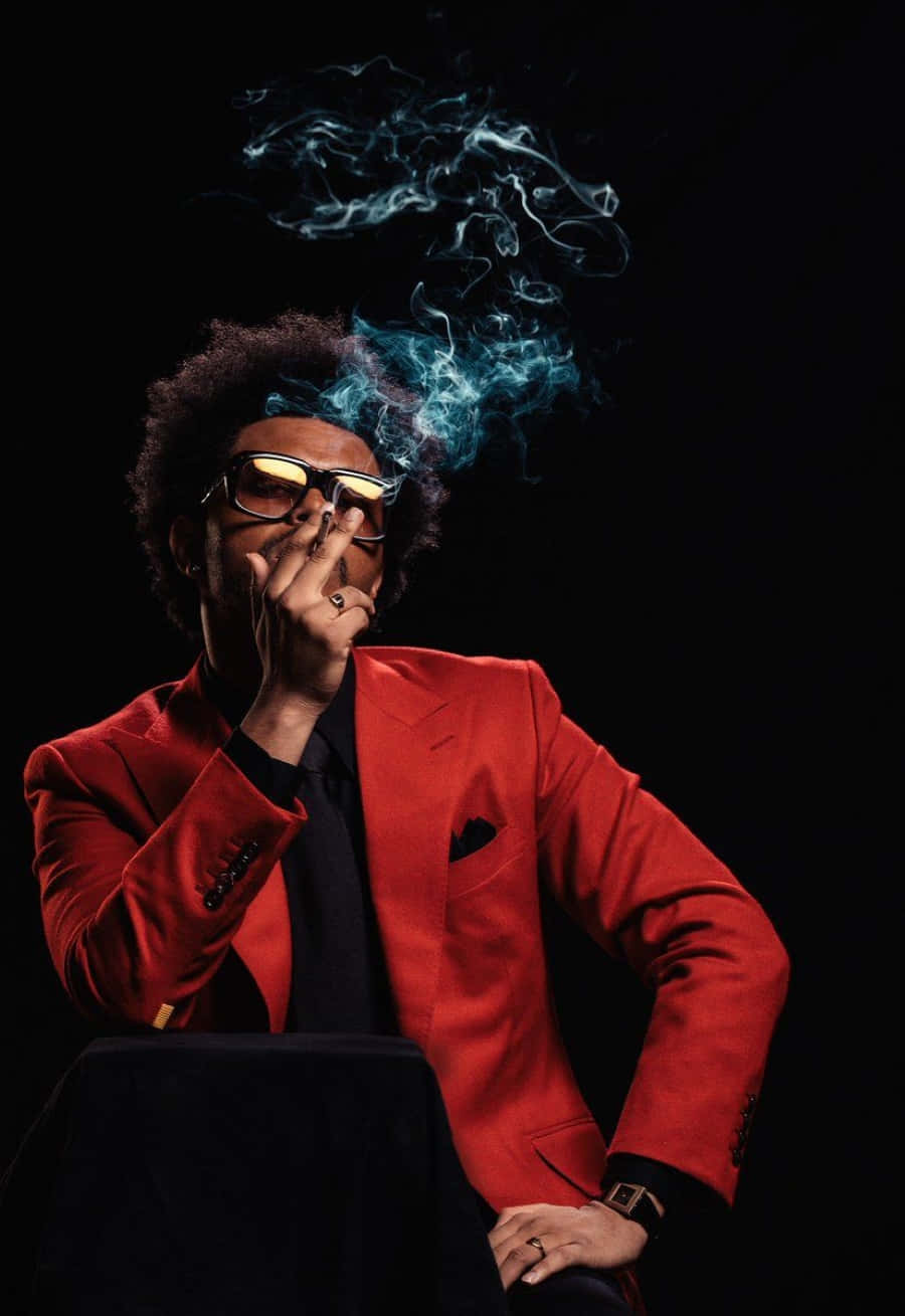 The Weeknd After Hours 桌布 壁紙 — 一位時尚男士身穿紅色西裝、戴著太陽眼鏡，在黑色背景前呼出發光的藍色煙霧。