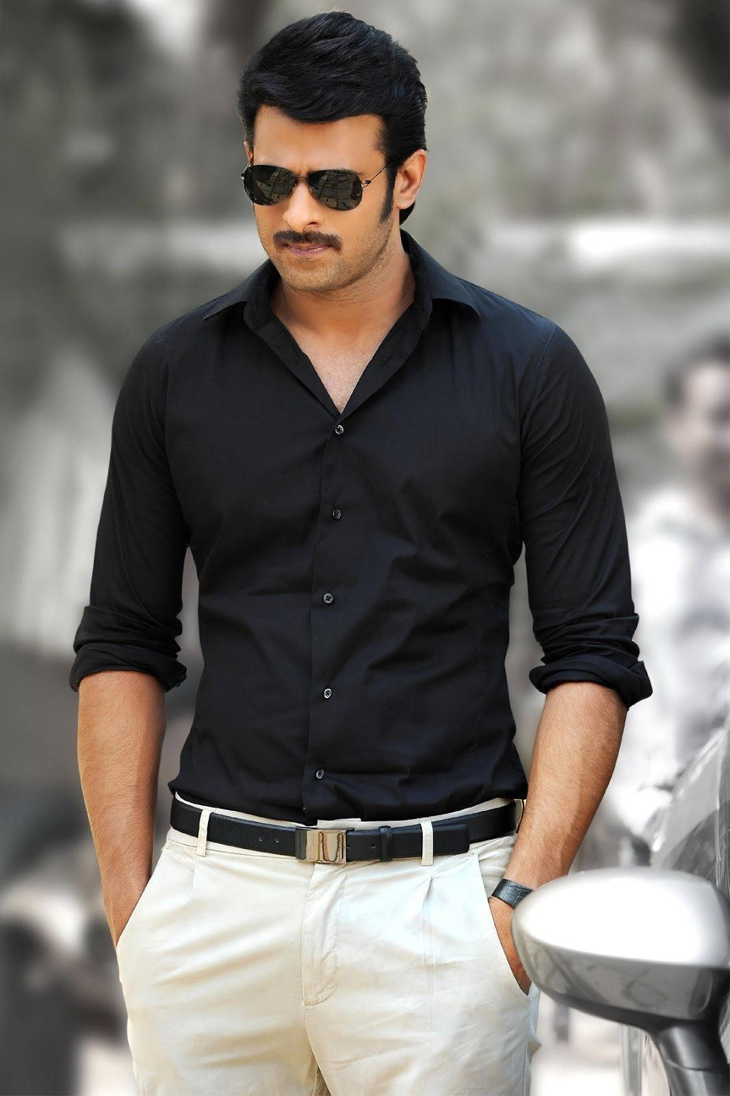 Prabhas Mirchi 桌布 壁紙 — 一位時尚男士身穿黑色襯衫和米色長褲，戴著太陽眼鏡自信地站在車旁。