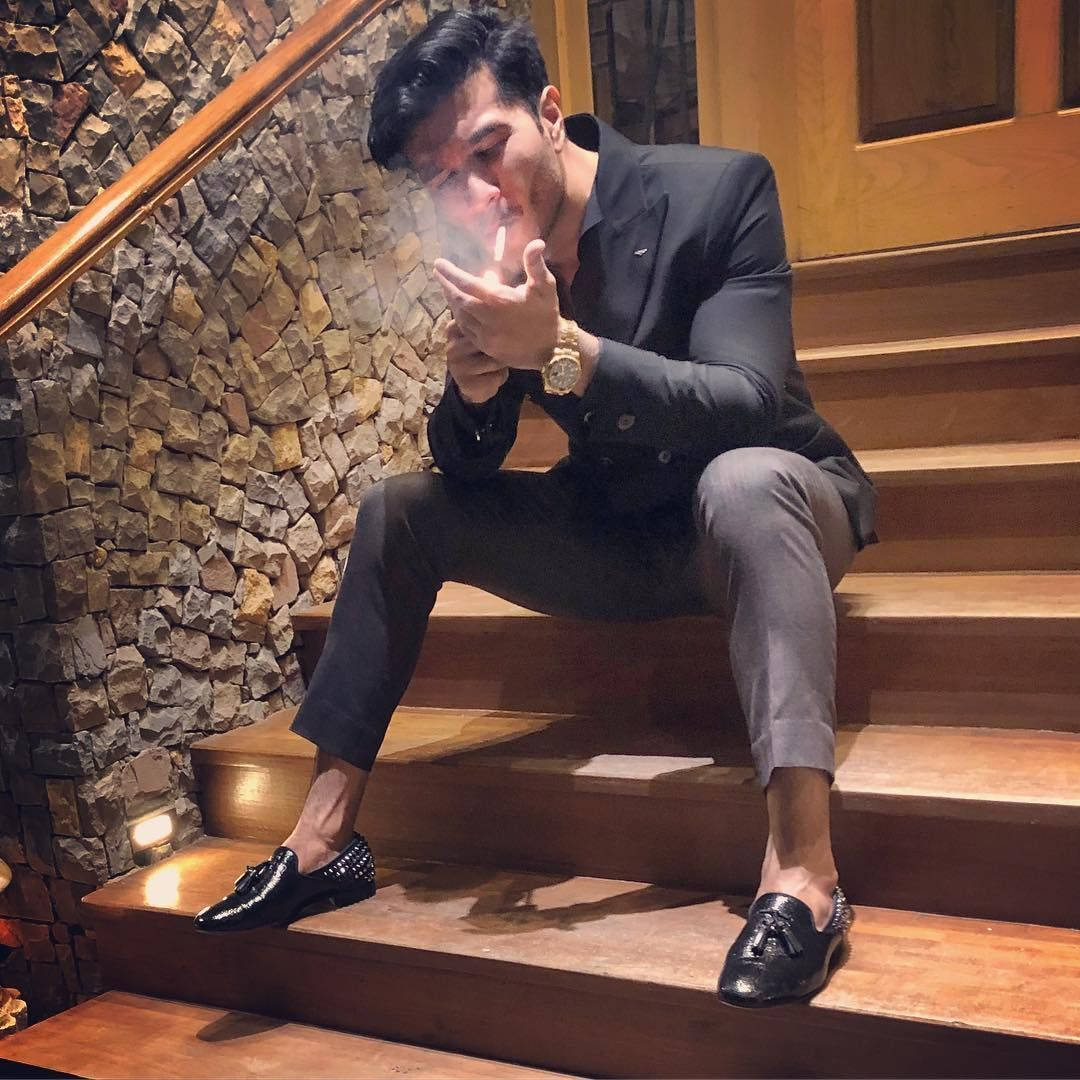 Feroz Khan 桌布 壁紙 — 一位時尚男士身穿黑色西裝坐在木樓梯上，對著質樸的石牆呼出煙霧。
