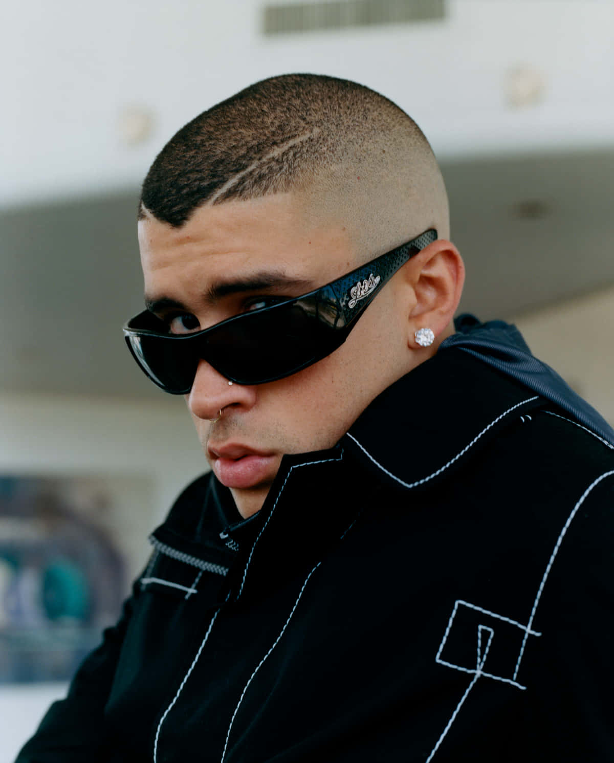Bad Bunny 桌布 壁紙 — 一位時尚的年輕男子，剃著光頭，戴著大膽的太陽眼鏡，在現代都市環境中凝視著鏡頭。