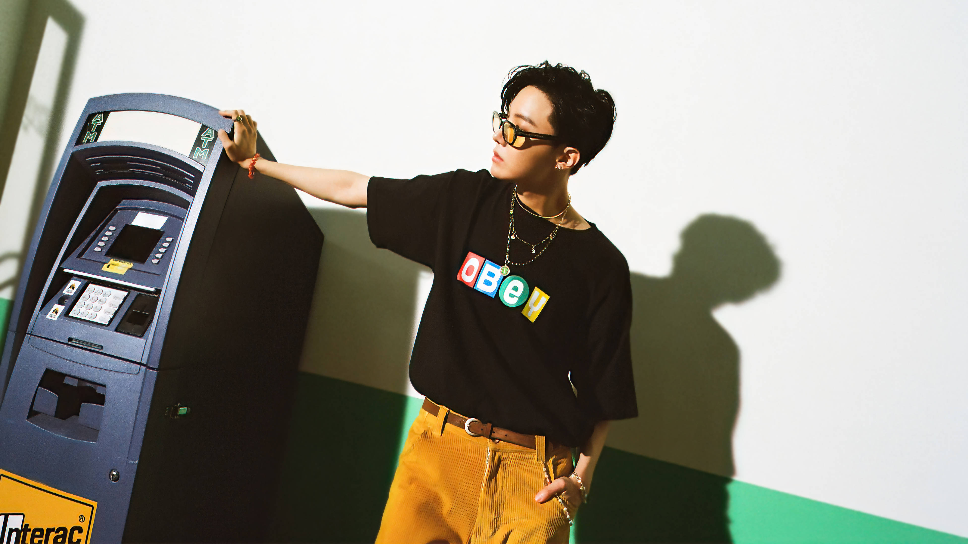 BTS J-hope Dynamite 壁紙 — 一位時尚的年輕男子身穿黑色OBEY T恤和芥末黃褲子，靠在一台藍色自動櫃員機上，在白綠相間的牆上投下大膽的影子。