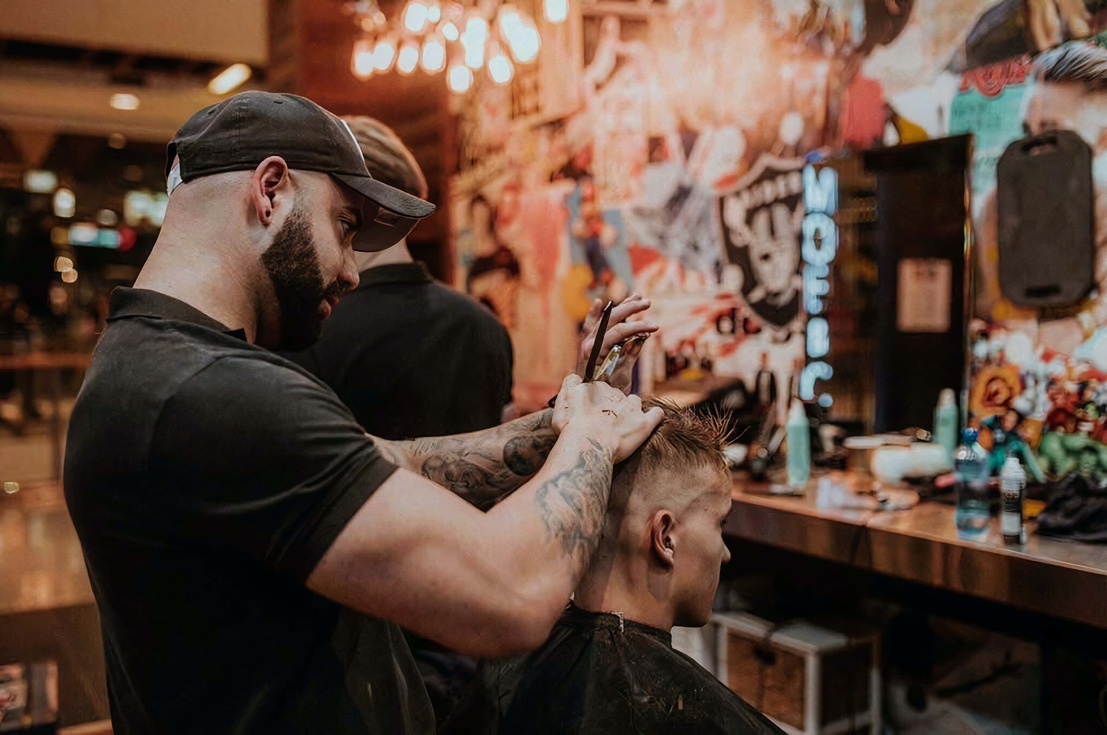Barber 桌布 壁紙 — 一位有紋身、戴帽子的理髮師在燈光溫暖、牆上有塗鴉的理髮店裡，小心翼翼地為一位年輕男子修剪頭髮。