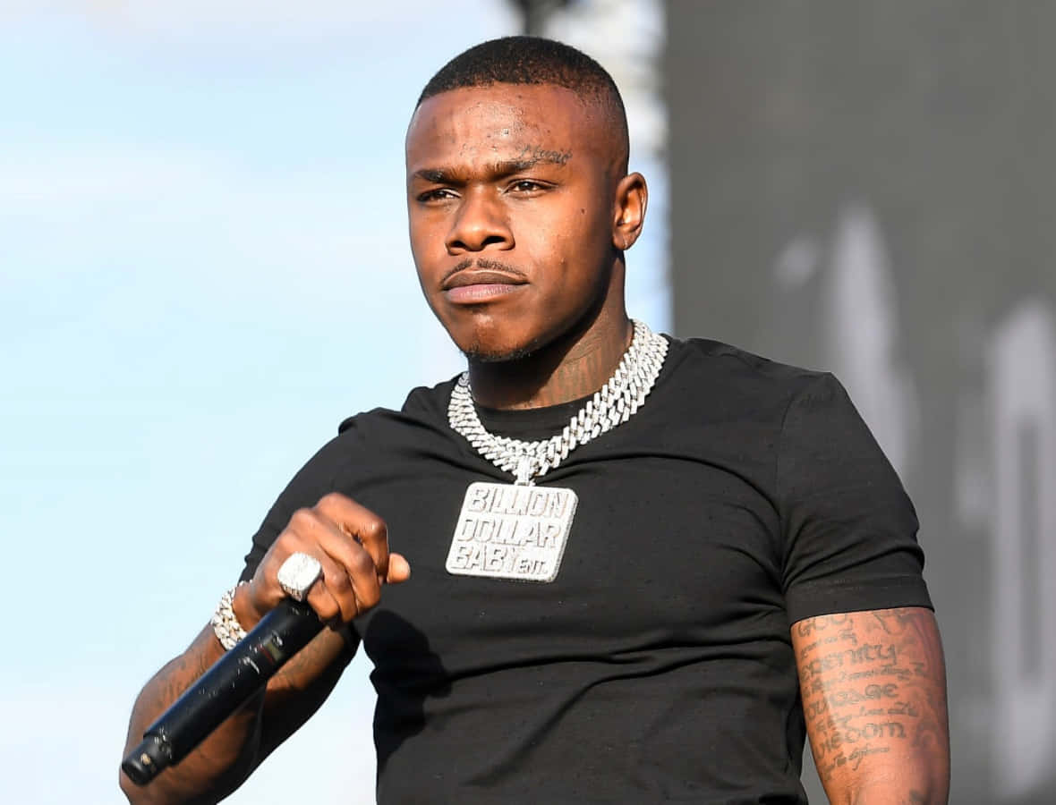 Dababy 桌布 壁紙 — 一位有紋身和粗鏈條項鍊的男性饒舌歌手在明亮的日光下戶外表演時手持麥克風。