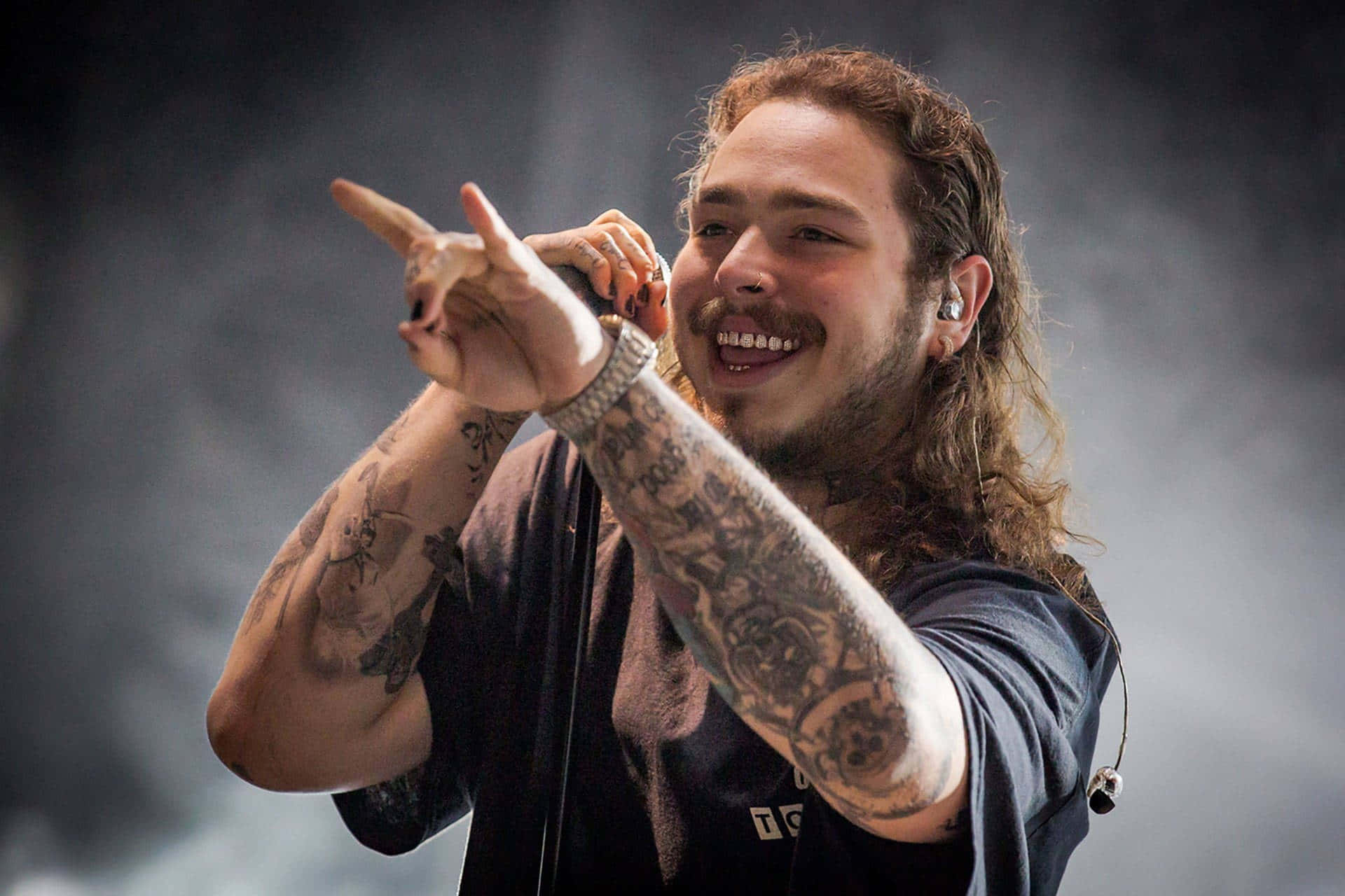 Post Malone 桌布 壁紙 — 一位有紋身嘅長髮音樂人喺煙霧彌漫嘅舞台上對住咪高峰唱歌，微笑並指住。