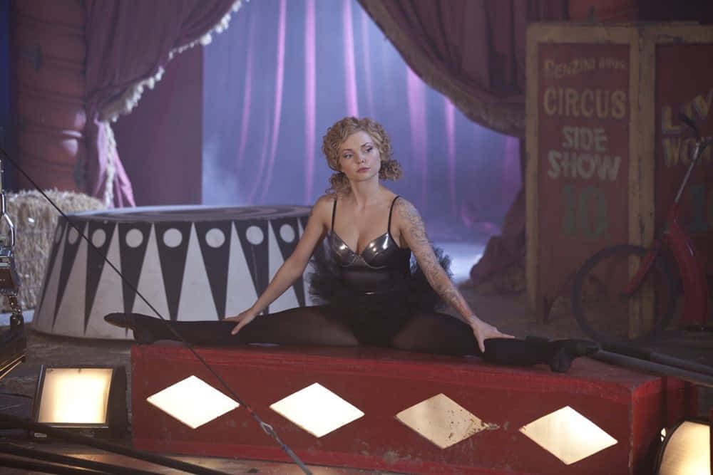 Izabella Miko 桌布 壁紙 — 一位有紋身的女士優雅地坐在紅色馬戲團平台上，紫色窗簾下，身後有「Circus Side Show」標誌。