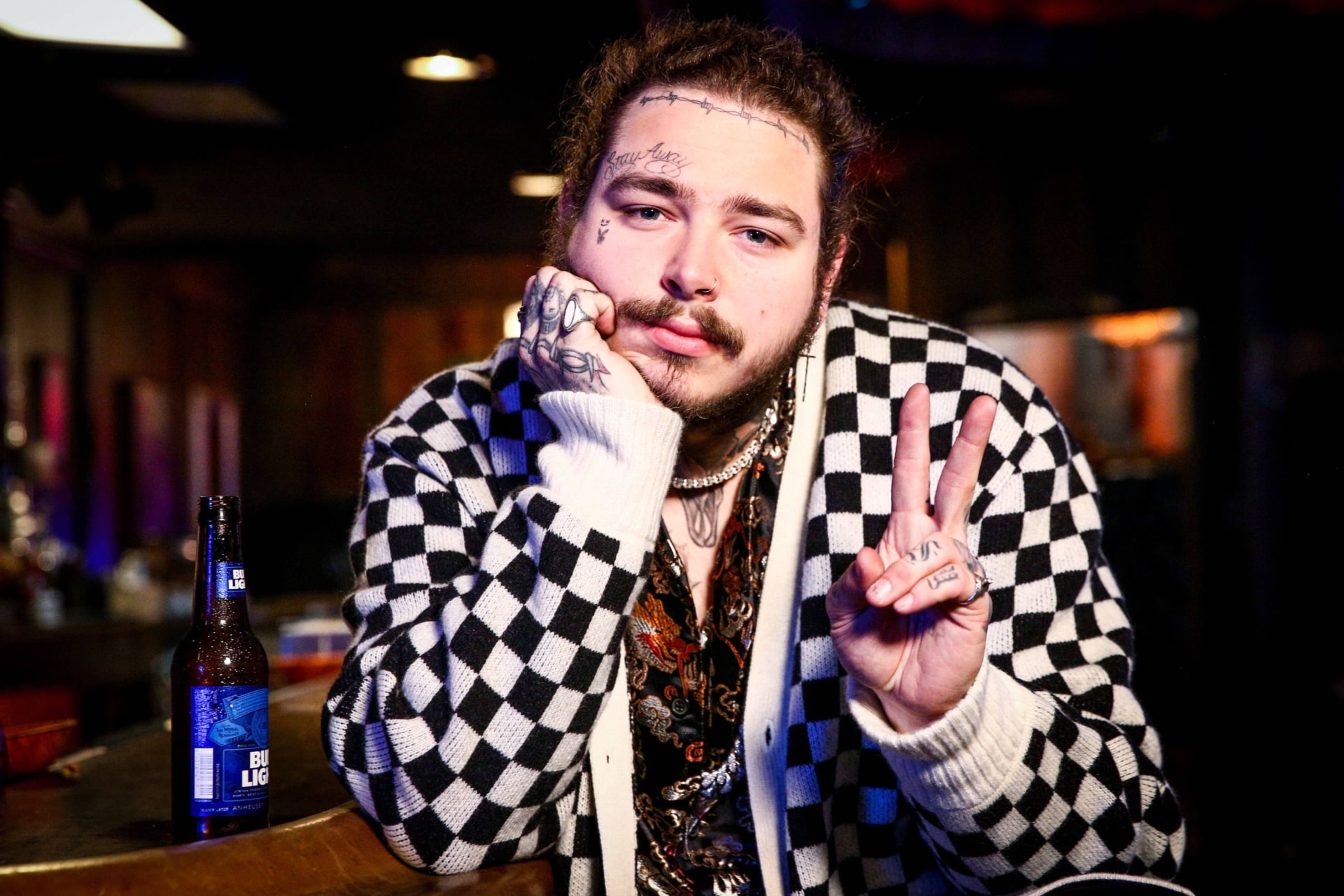 Post Malone Footsies Dive Bar 壁紙 — 一位有紋身的男士穿著黑白格仔毛衣，在燈光昏暗的酒吧裡托腮做出和平手勢。