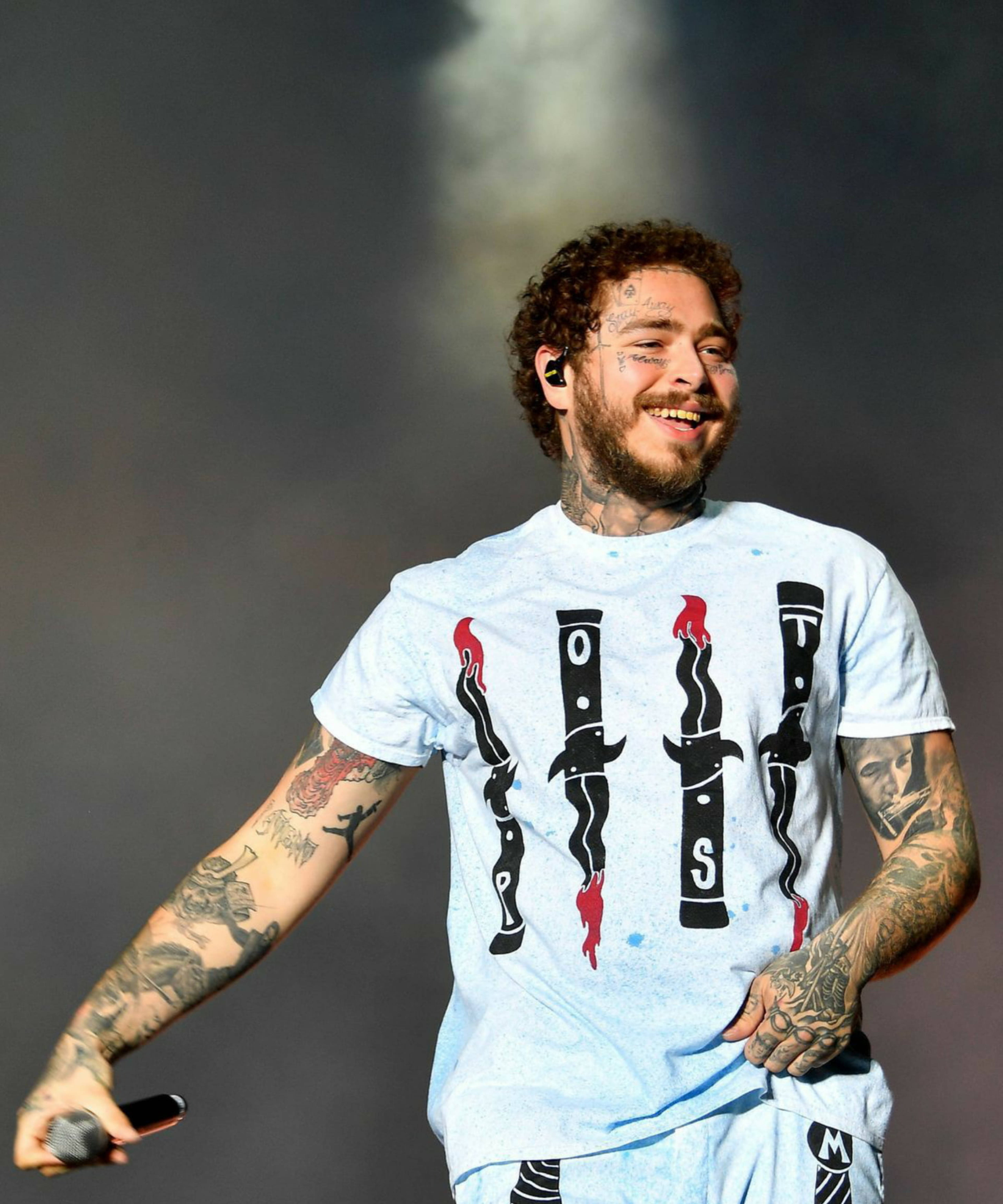 Post Malone 2019 Sziget Festival 壁紙 — 一位有紋身的男性表演者在煙霧彌漫的音樂會舞台上笑著拿著麥克風。