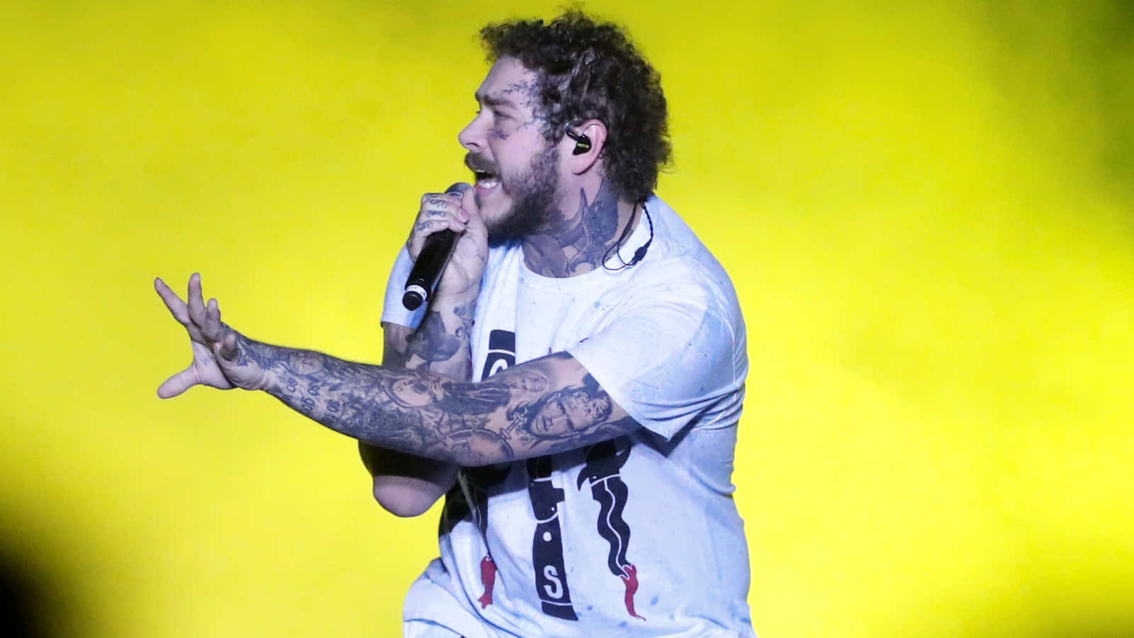 Post Malone 桌布 壁紙 — 一位有紋身的男歌手在充滿活力的黃色背景下於舞台上熱情演出。