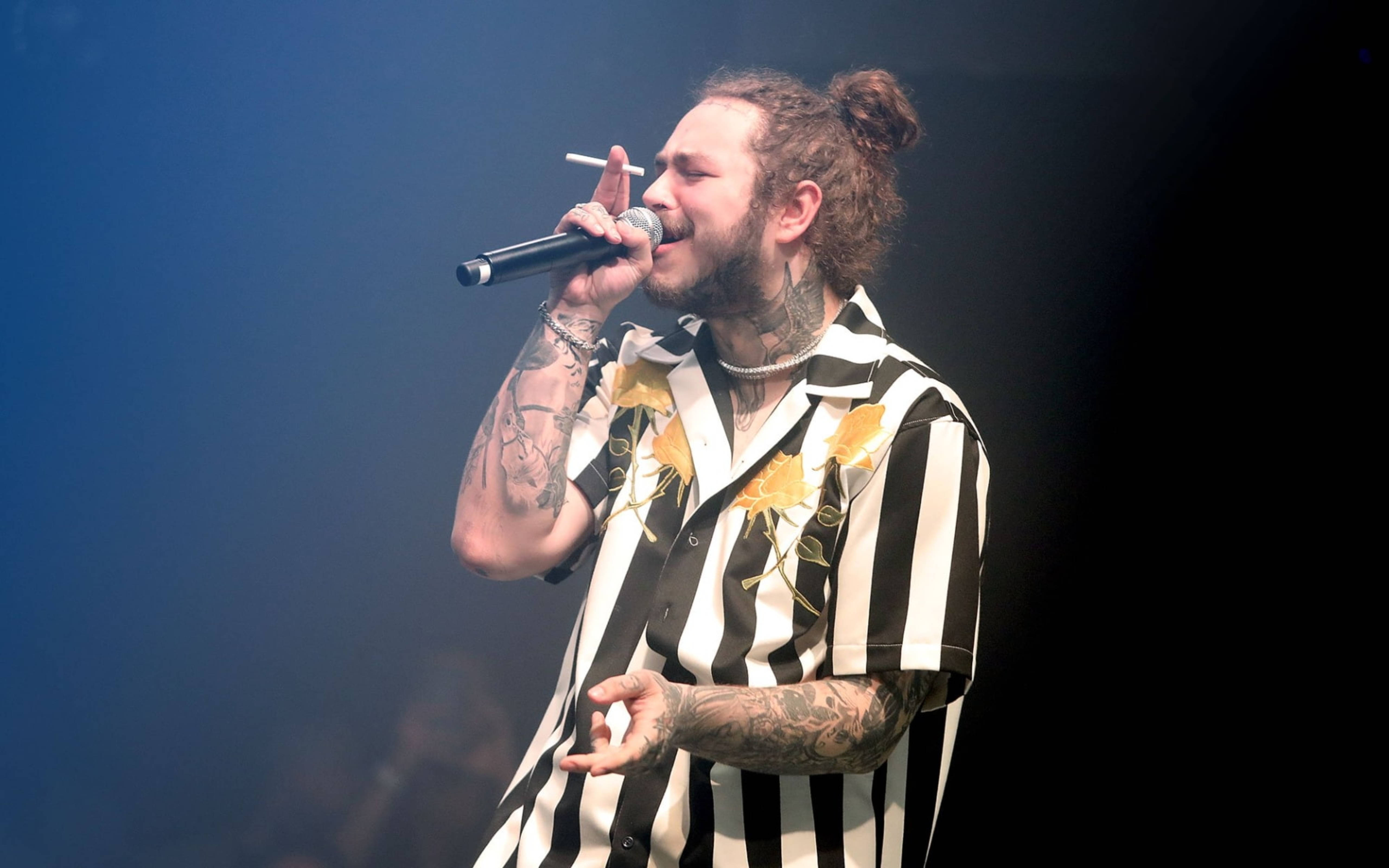 Post Malone Concert 壁紙 — 一位有紋身的男歌手紮著髮髻，在藍色舞台燈光下拿著麥克風和香煙表演。
