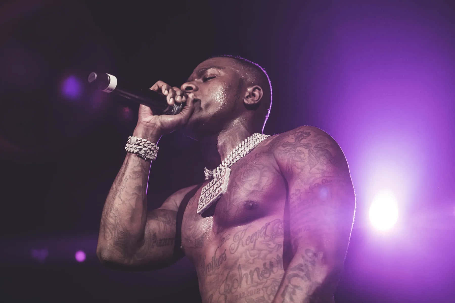 Dababy 桌布 壁紙 — 一位有紋身的男饒舌歌手在充滿活力的紫色舞台燈光下，對著麥克風熱情表演。