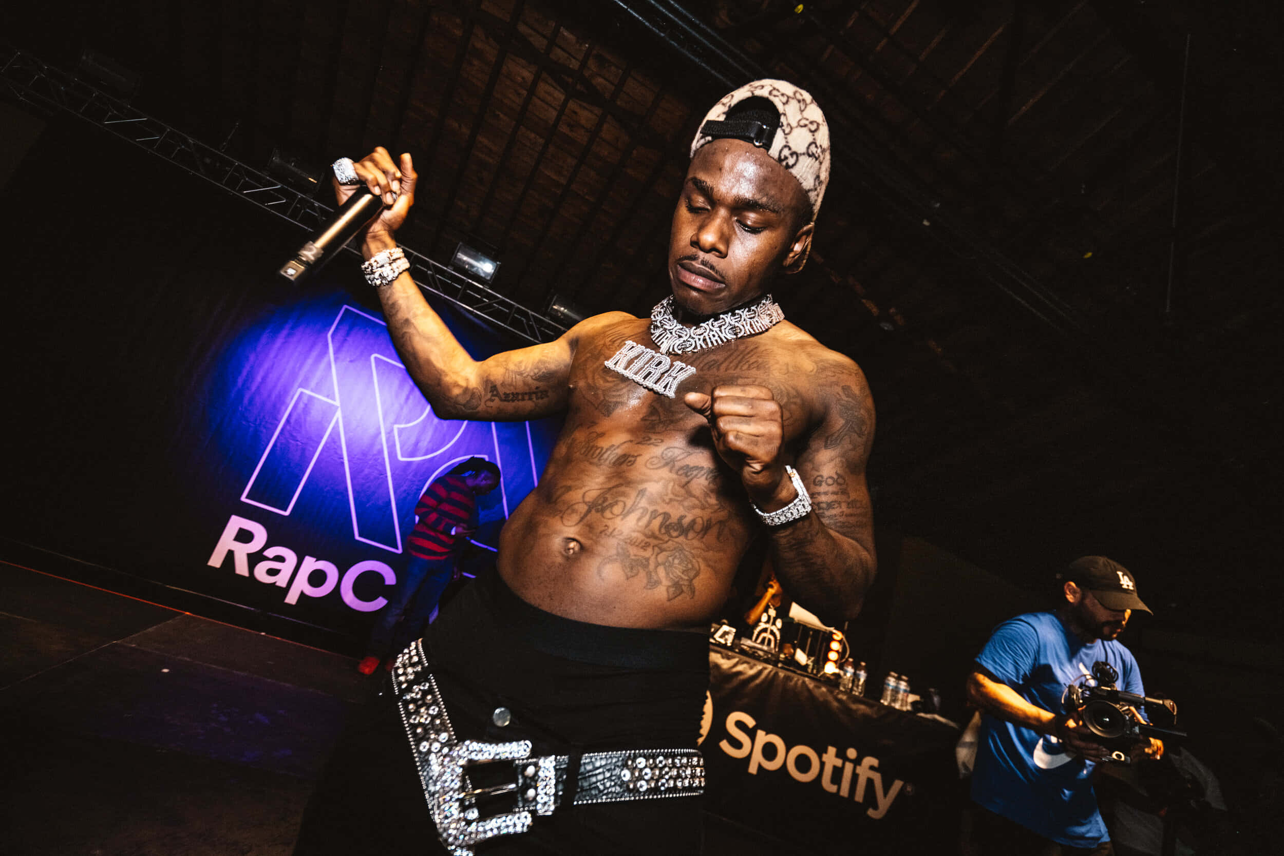 Dababy 桌布 壁紙 — 一位有紋身的說唱歌手在紫色聚光燈下拿著麥克風表演，身後有一位DJ。