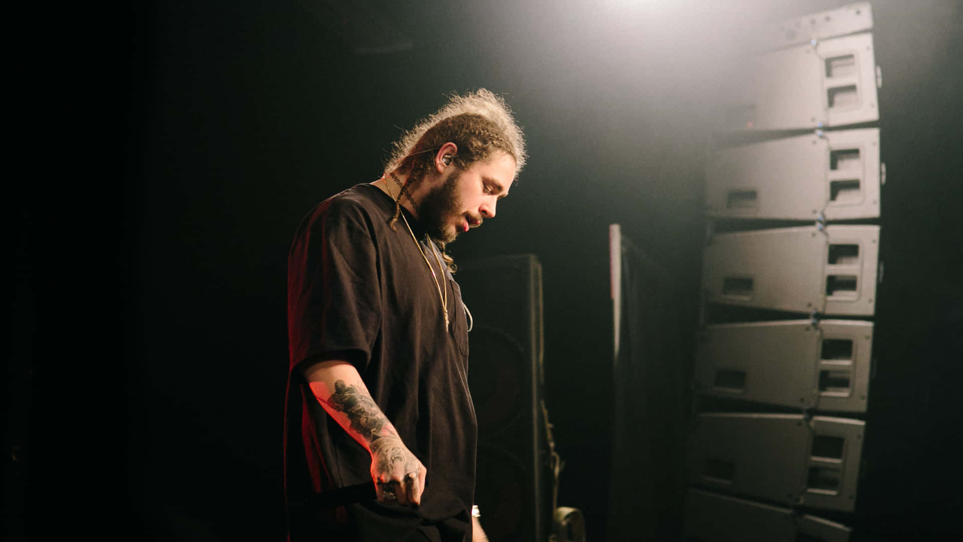 Post Malone 桌布 壁紙 — 一位有紋身的音樂家，捲髮，在昏暗的舞台燈光下站在堆疊的音箱旁，低頭專注。