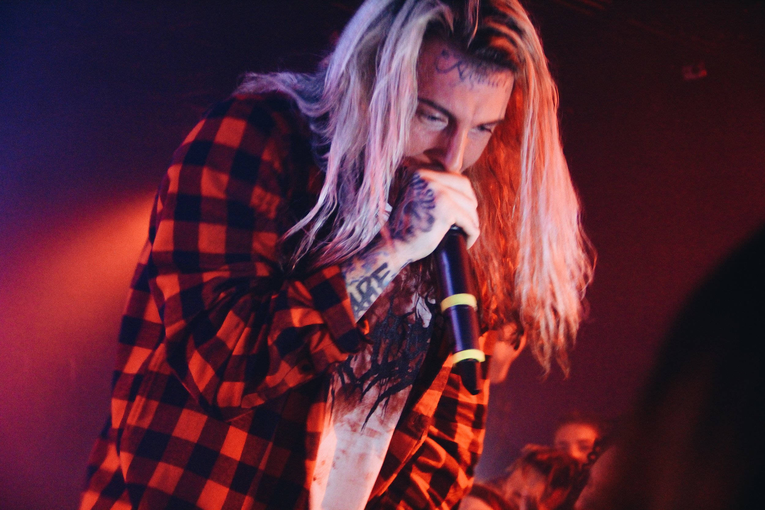 Ghostemane 桌布 壁紙 — 一位有紋身的音樂家，長長的金髮，在紅色舞台燈光下熱情地對著麥克風唱歌。