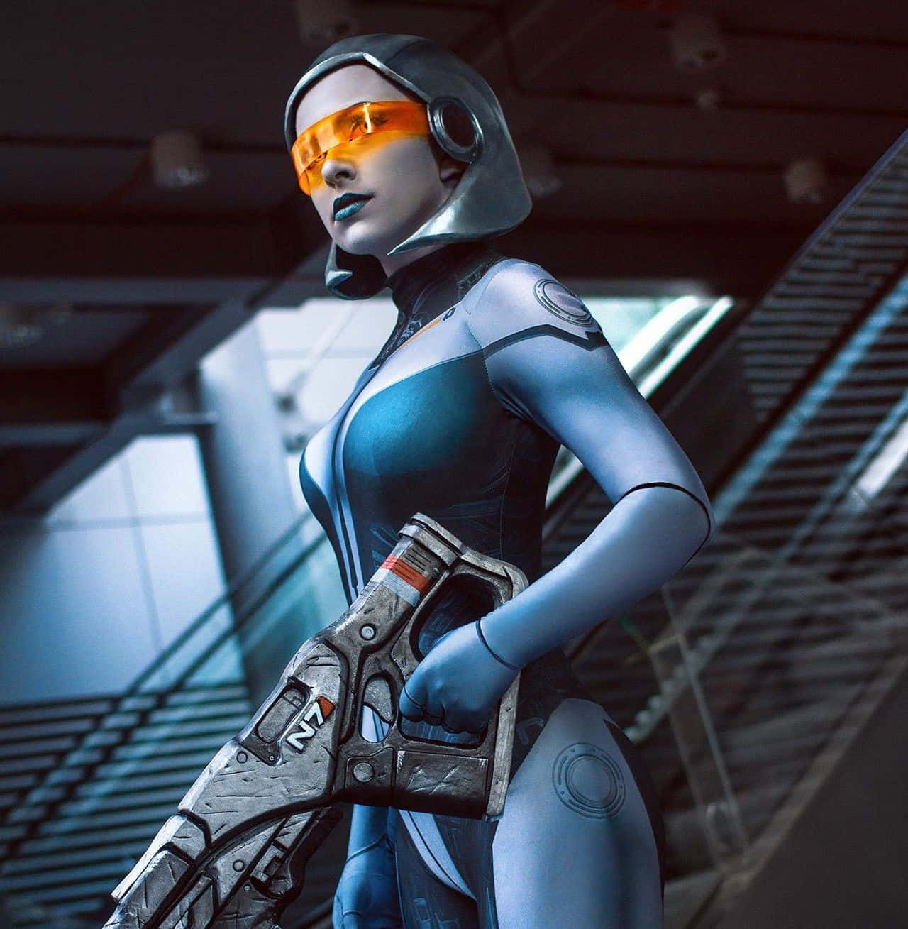 Mass Effect Edi 桌布 壁紙 — 一位未來感的女戰士，身穿流線型藍色裝甲和發光橙色面罩，在金屬走廊中手持高科技武器。