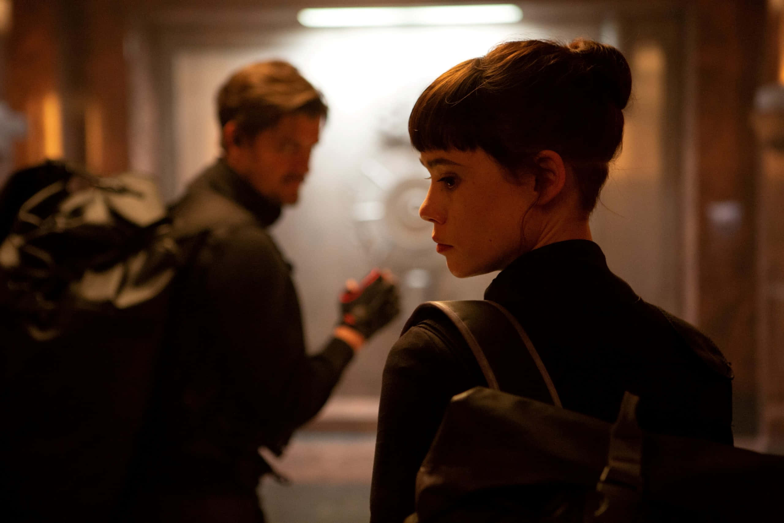 Astrid Berges Frisbey 桌布 壁紙 — 一位束著髮髻、背著背包的女子在昏暗的走廊中轉頭，身後有一位背著背包的男子走過。