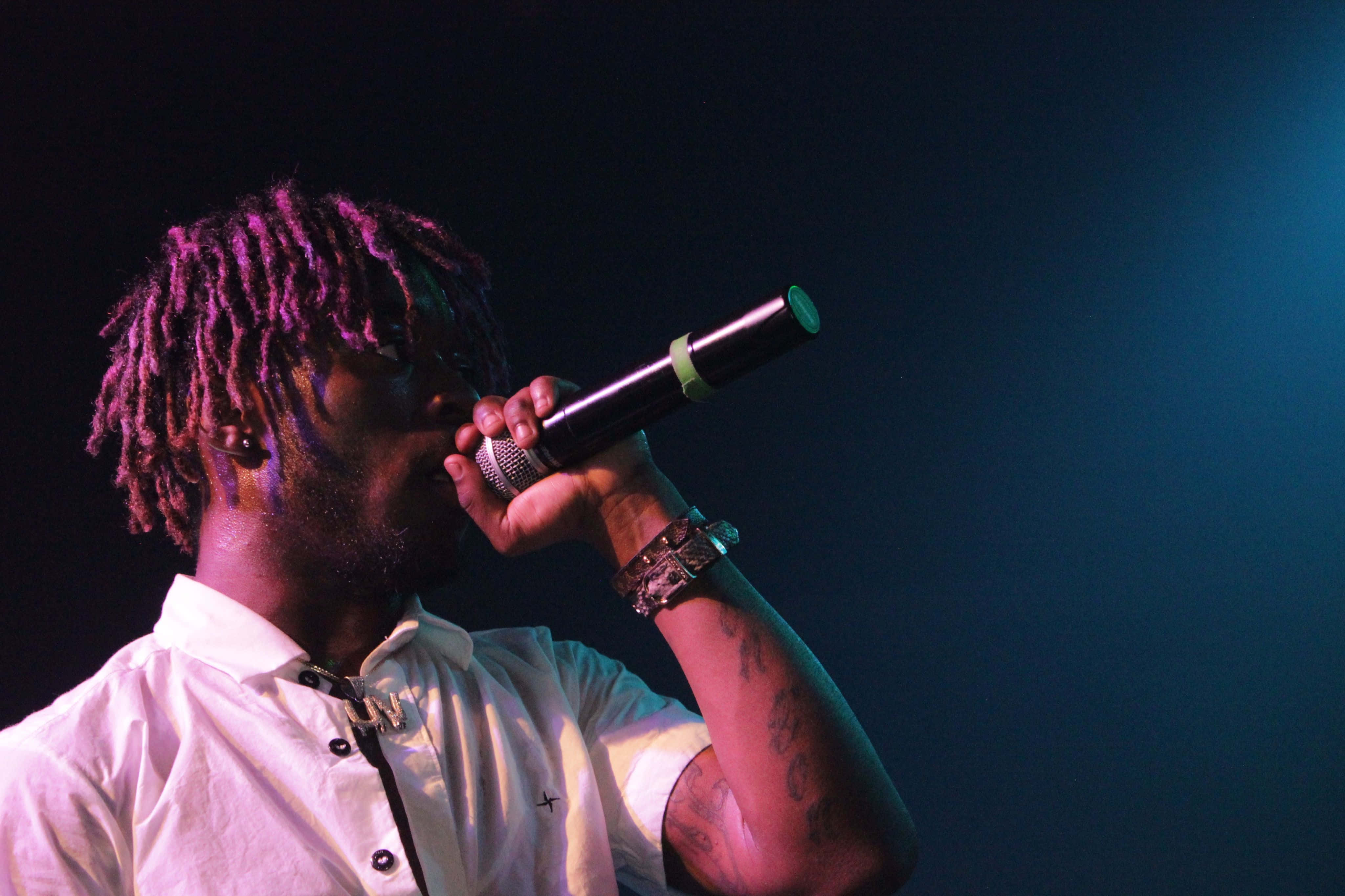 Lil Uzi Vert 桌布 壁紙 — 一位染有紫色辮子的說唱歌手，身穿白襯衫，在藍色舞台燈光下對著麥克風唱歌。