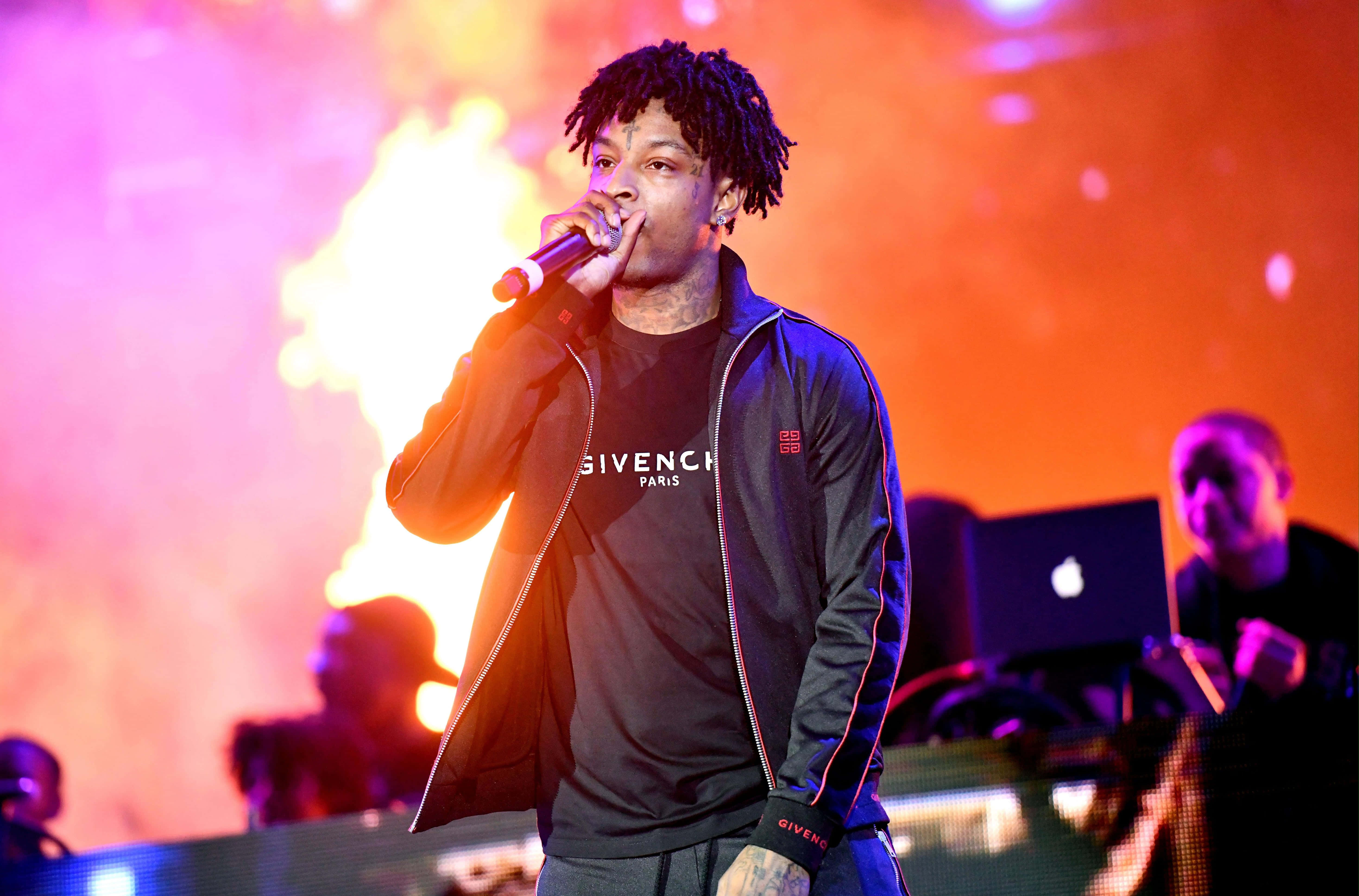 21 Savage 壁紙 — 一位梳著辮子的說唱歌手在熾熱的橙色和粉紅色燈光下拿著麥克風在舞台上表演。