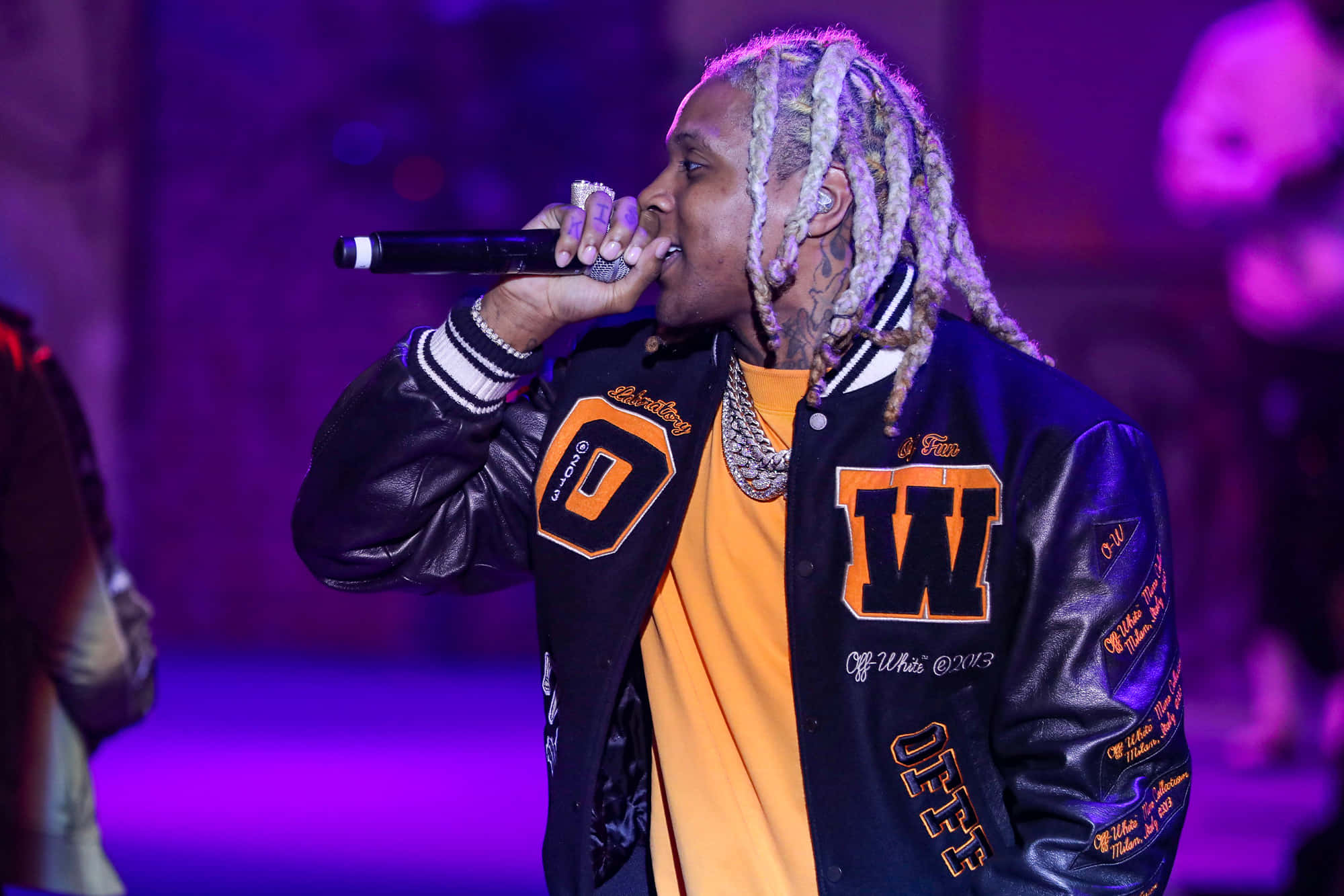 lil durk 桌布 壁紙 — 一位梳著辮子的說唱歌手在紫色燈光下穿著帶有橙色字母的黑色校隊夾克在舞台上表演。